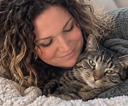 A woman with curly hair lying on a bed beside a tabby cat, both appearing relaxed and content.