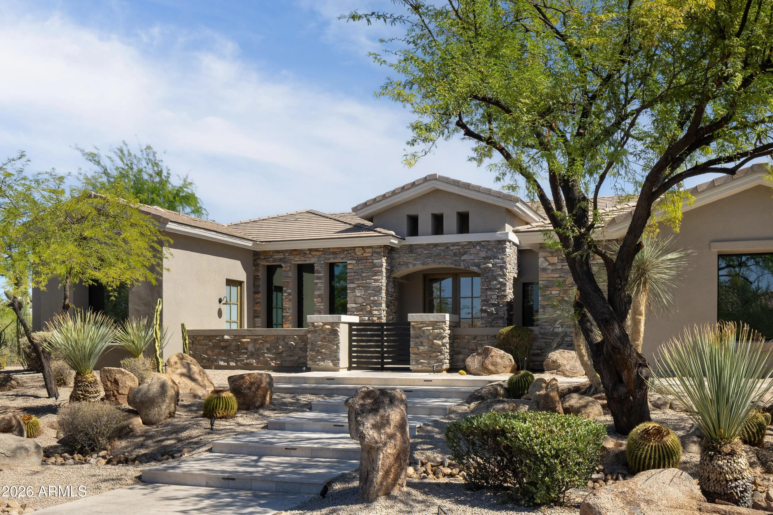 Front view of a modern house with stone facade, surrounded by desert landscaping including cacti, rocks, and drought-resistant plants. A large tree with green leaves is in the foreground, and the sky is partly cloudy.