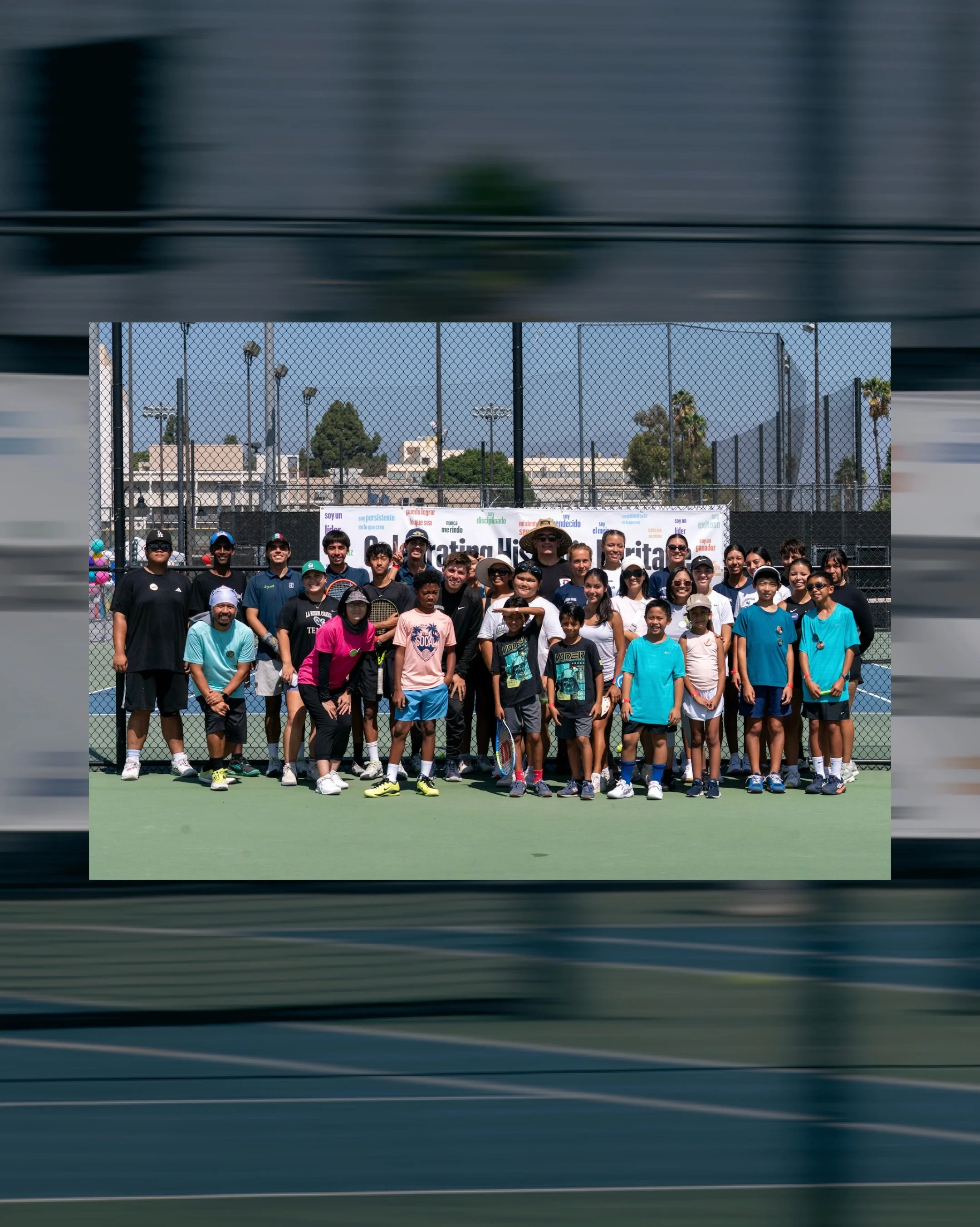 Group of children and adults standing in front of a tennis court with a large banner and high chain-link fence. Some children hold tennis rackets, and most people are wearing casual sports clothing and hats.