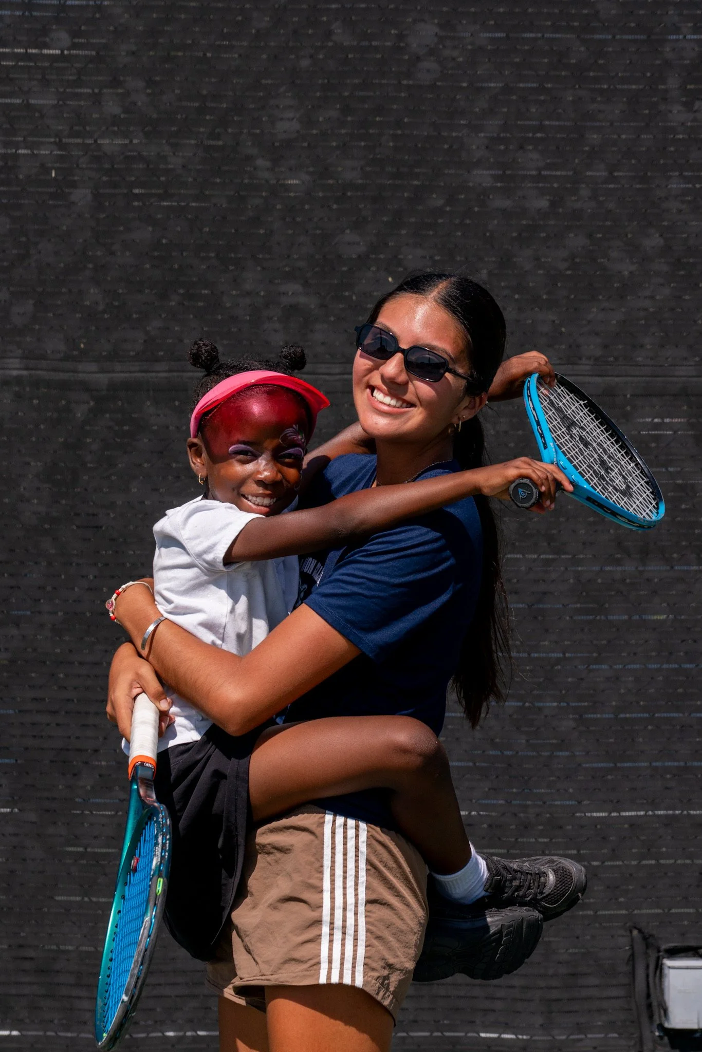 A woman with dark hair wearing sunglasses and a navy blue shirt is holding a young girl with a pink headband, colorful face paint, and sporting a white shirt and black shorts. They are on a tennis court with a black fence in the background. The woman is smiling as she lifts the girl who is also smiling.