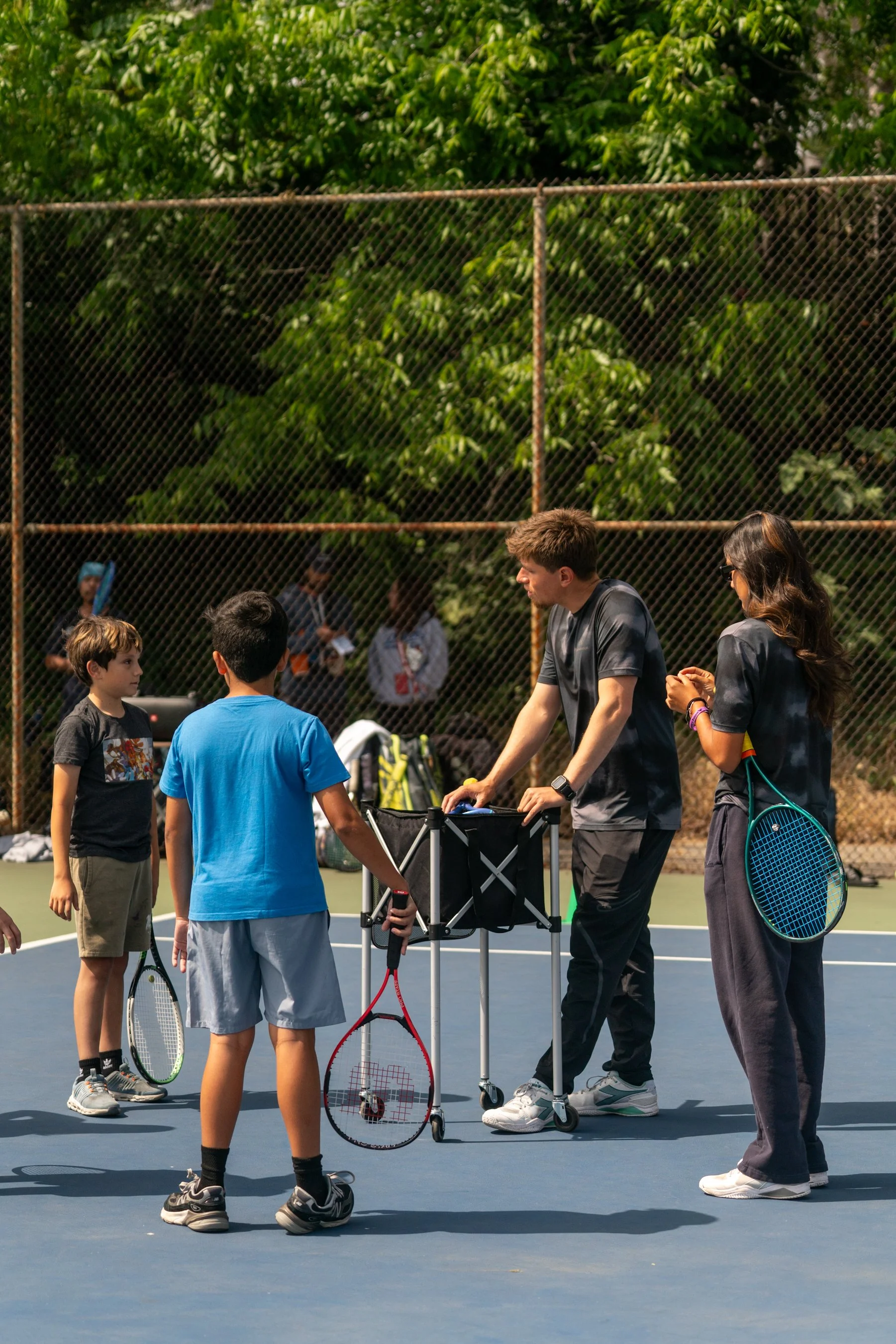 Group of children and young adults on a tennis court, some holding tennis rackets, with a man setting up tennis balls and equipment, and lush green trees in the background.