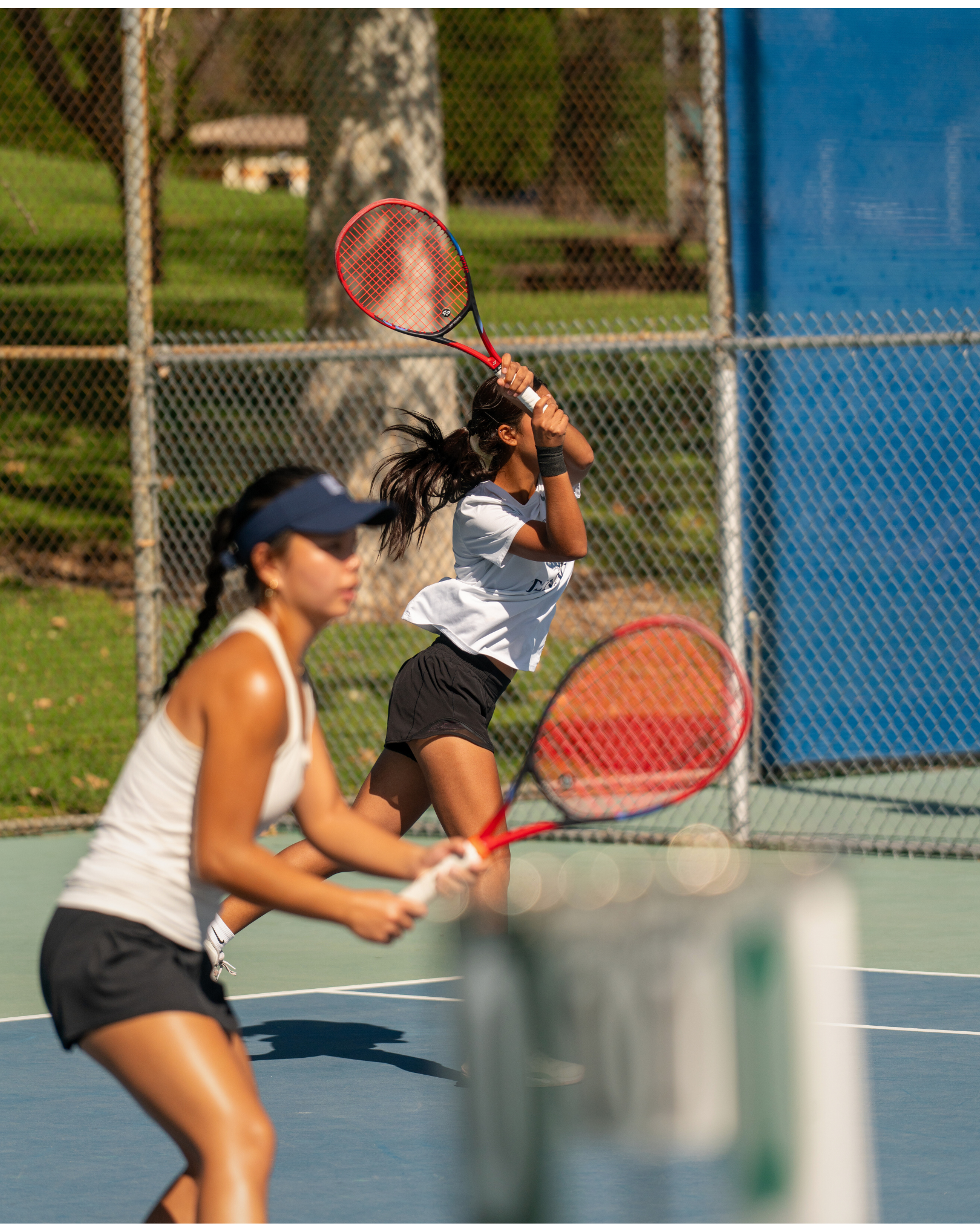 Two young women playing tennis on an outdoor court during daytime, with a chain-link fence and trees in the background.