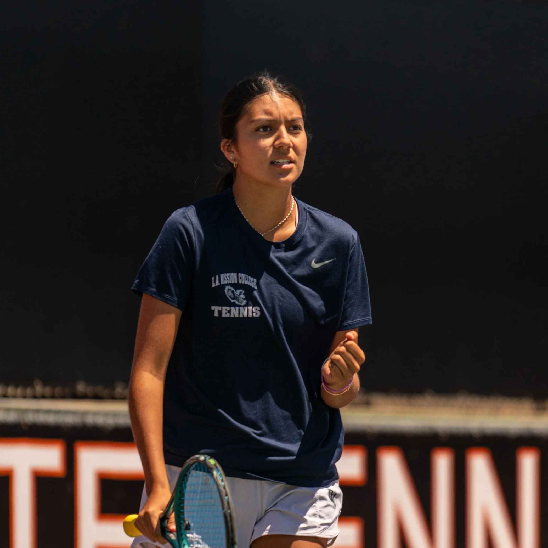 Young female tennis player on court, wearing a navy T-shirt with college logo, holding tennis racket, in a moment of victory or celebration.