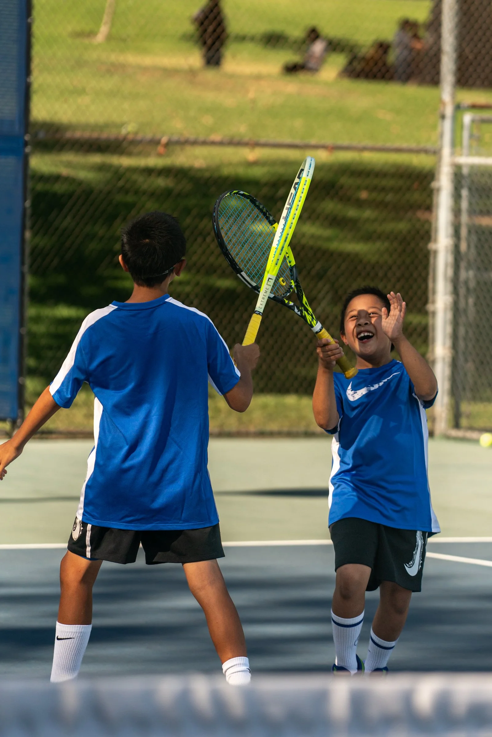 Two boys in blue sports jerseys and shorts playing tennis on an outdoor court, with two other children sitting behind a wire fence in the background.