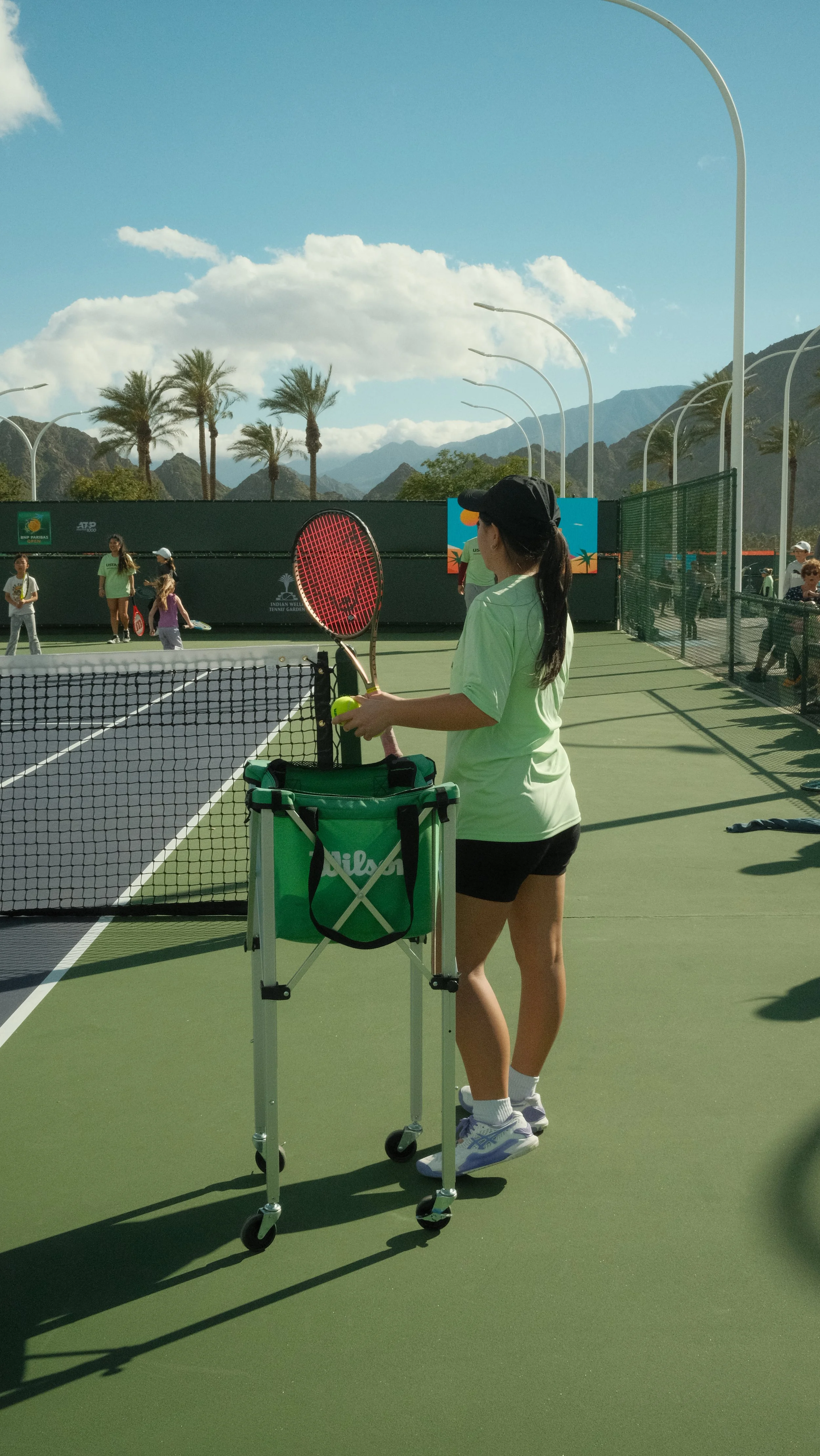 A woman holding a tennis racket and tennis ball standing on a tennis court with a tennis ball cart nearby. Several other people are on the court, and mountains, palm trees, and a blue sky with clouds are in the background.