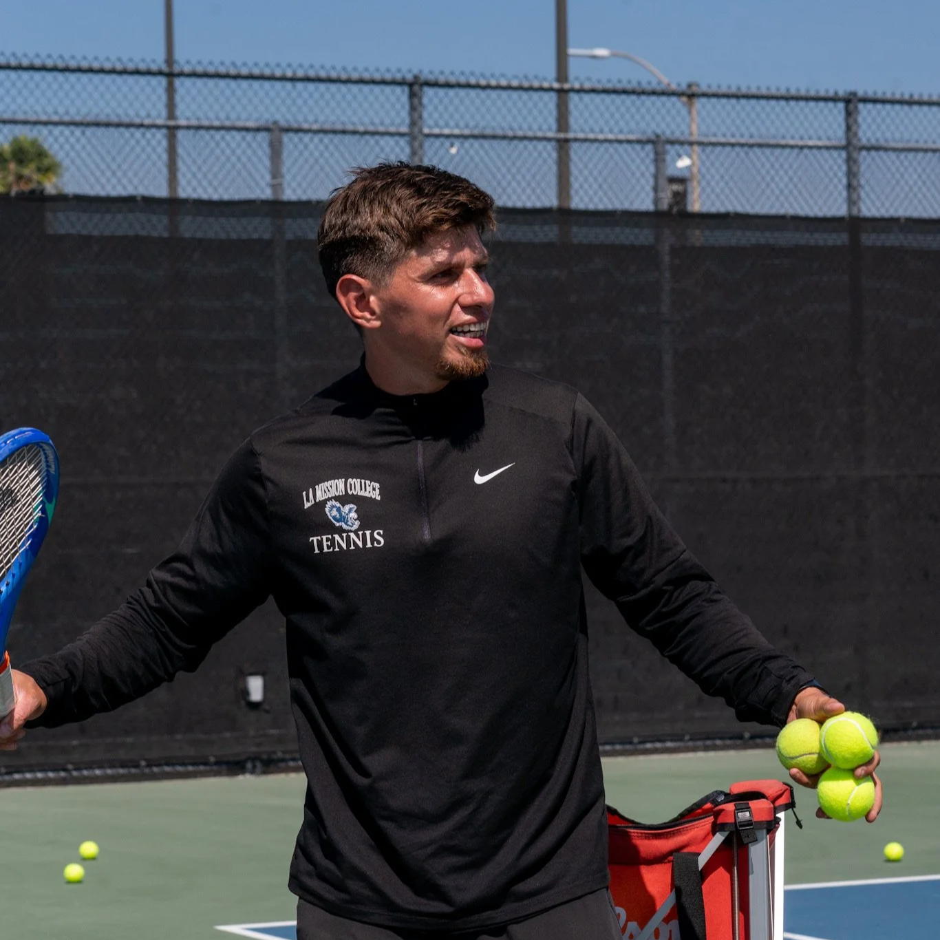A young male tennis player holding four tennis balls in his right hand and a racquet in his left hand, standing on a tennis court. He's wearing a black athletic jacket with the words 'La Mission College Tennis' and a lion logo on it. There are tennis balls on the court, and a black fence surrounds the court under a blue sky.