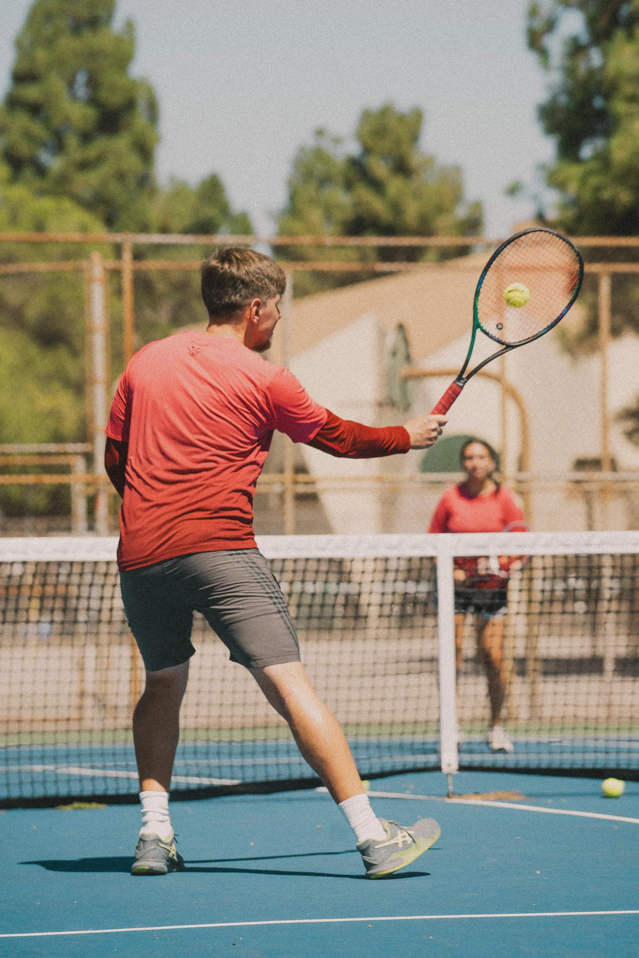 A man playing tennis on an outdoor court, hitting a tennis ball with a racket, with a woman watching in the background.