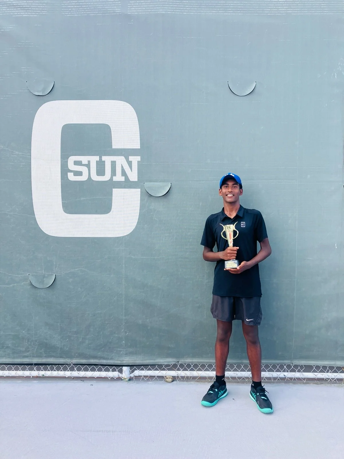 A young man standing on a tennis court holding a trophy, with a large sign that reads 'C SUN' in the background.