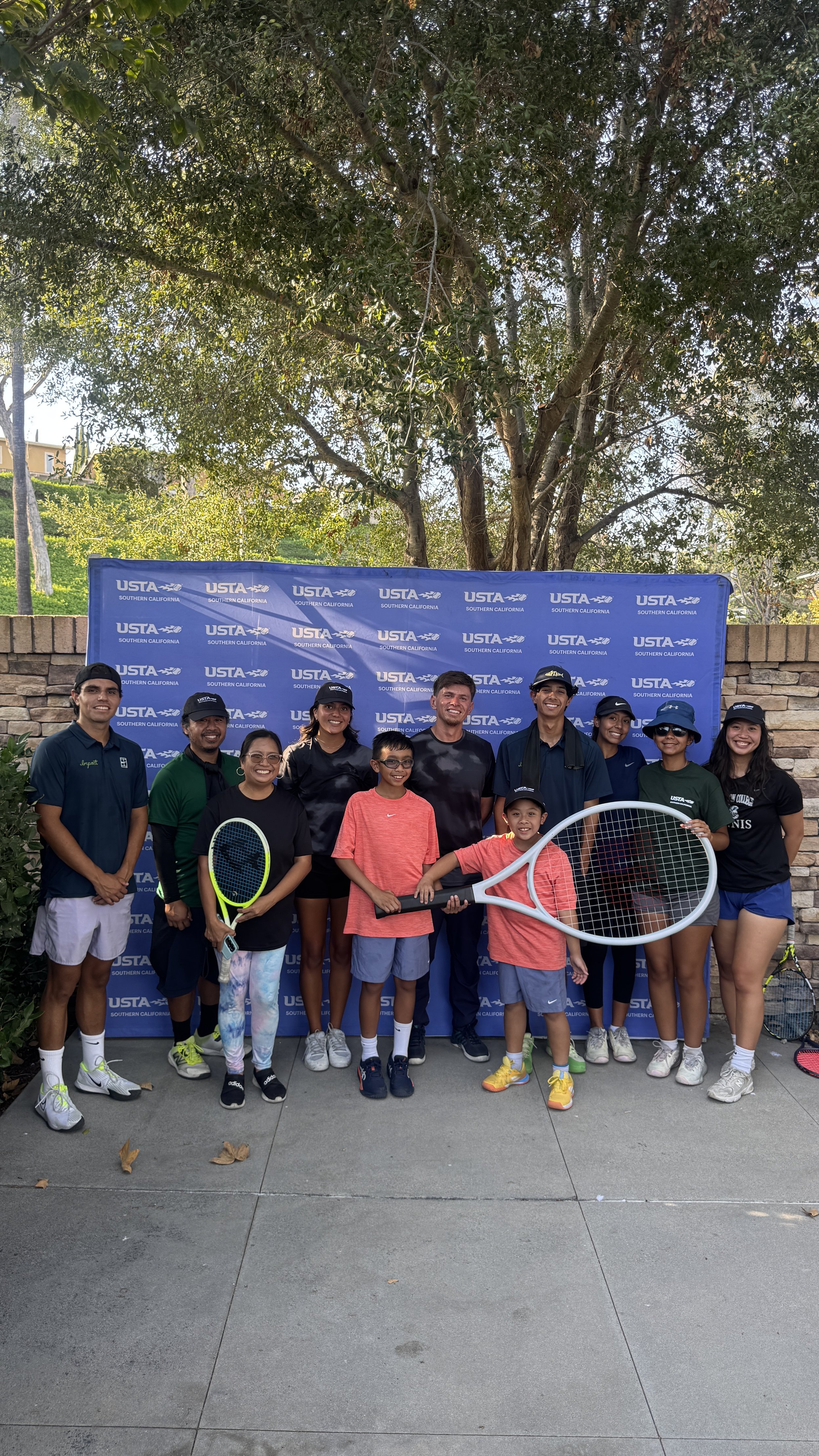 A group of 12 people, including children and adults, standing outdoors in front of a USTA Southern California backdrop, holding tennis rackets, some wearing sportswear and caps, smiling for a group photo.