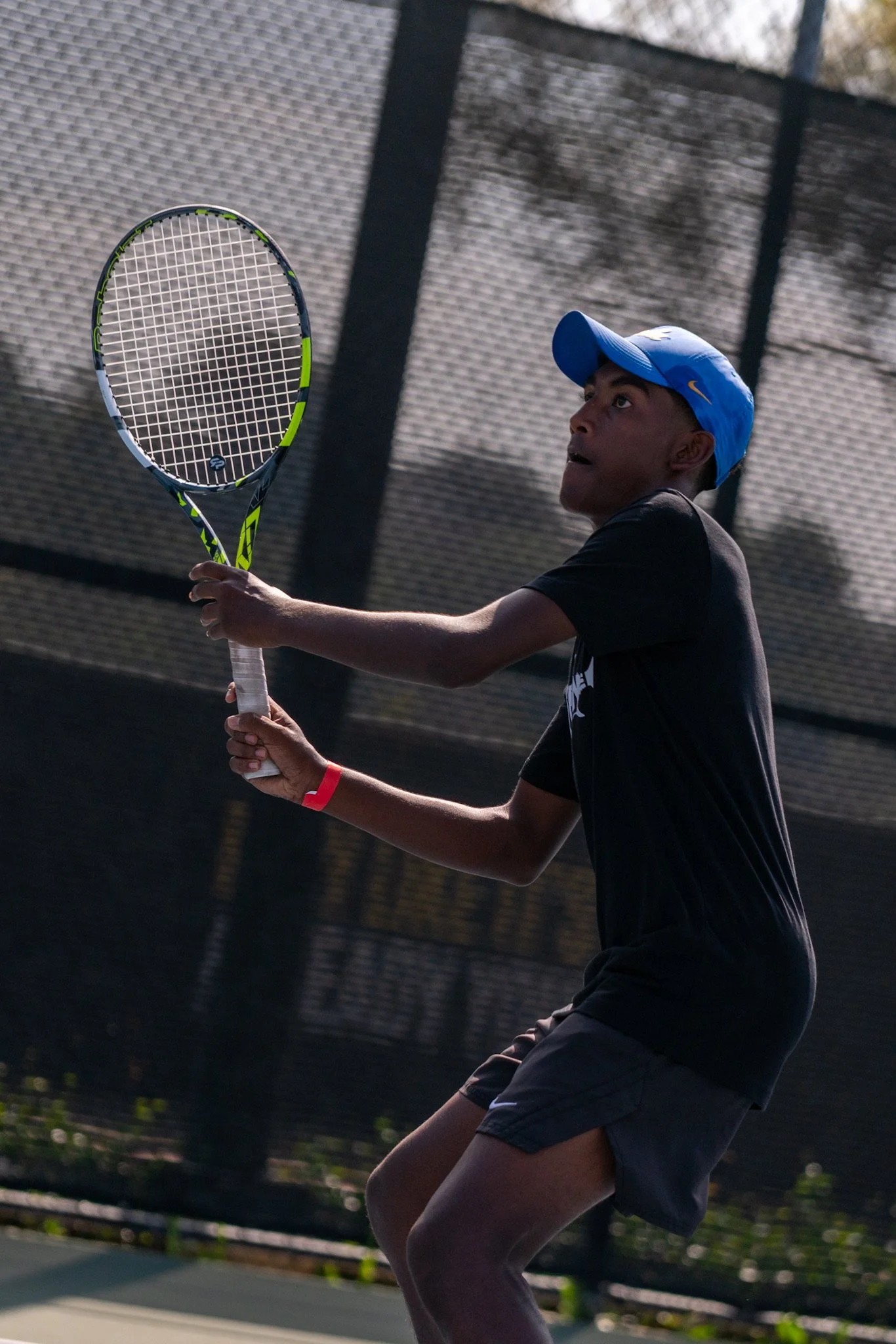 Young player on an outdoor tennis court, preparing to hit a tennis ball with his racket. The player is wearing a blue cap and dark athletic clothing.