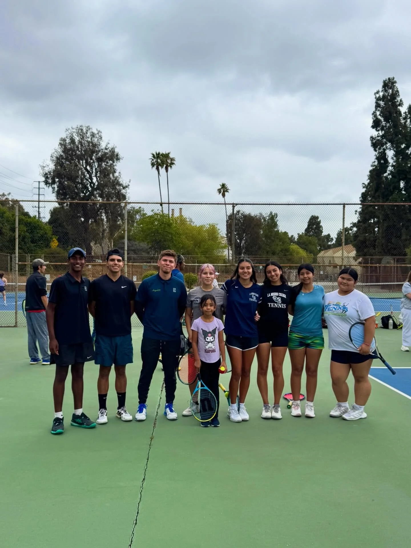 Group of young men and women standing together on a tennis court, holding tennis rackets, with trees and a fenced outdoor tennis court background under cloudy skies.