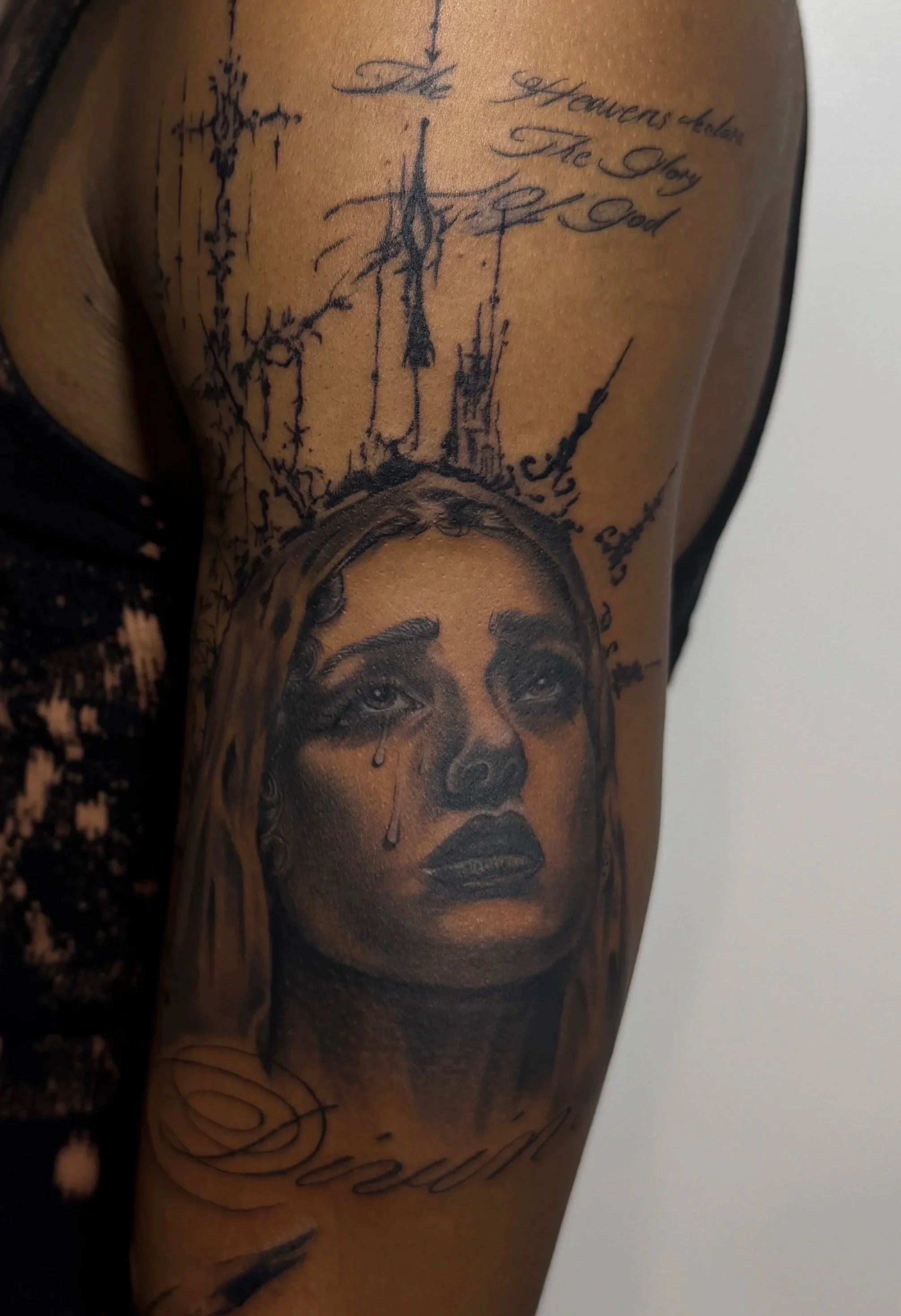 Close-up of a person's upper arm tattoo featuring a portrait of a woman with tears, a crown, and handwritten text above.