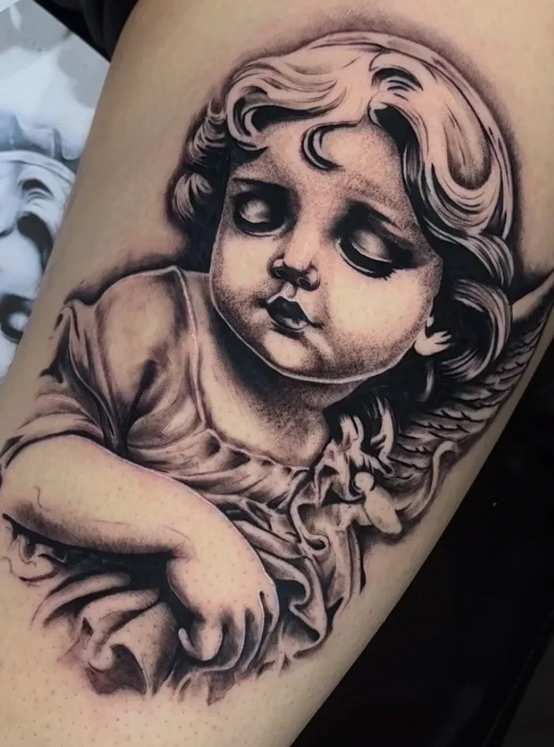 A black and gray tattoo of a child with a doll-like cherub face, featuring detailed hair, closed eyes, and a slight smile, resembling a classical style.