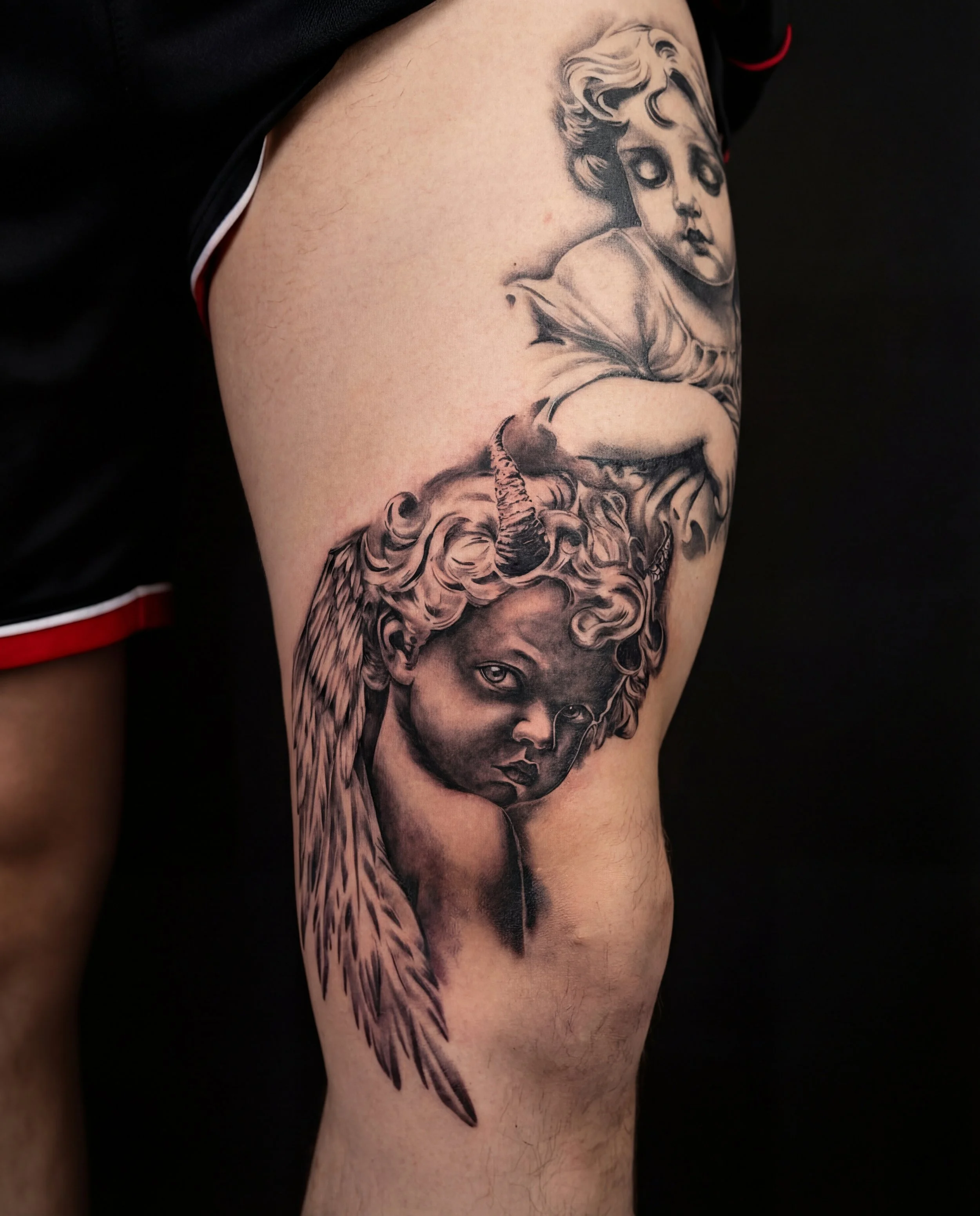 Black and gray tattoo on person's upper thigh of a dark cherub, accompanied by healed, light cherub.