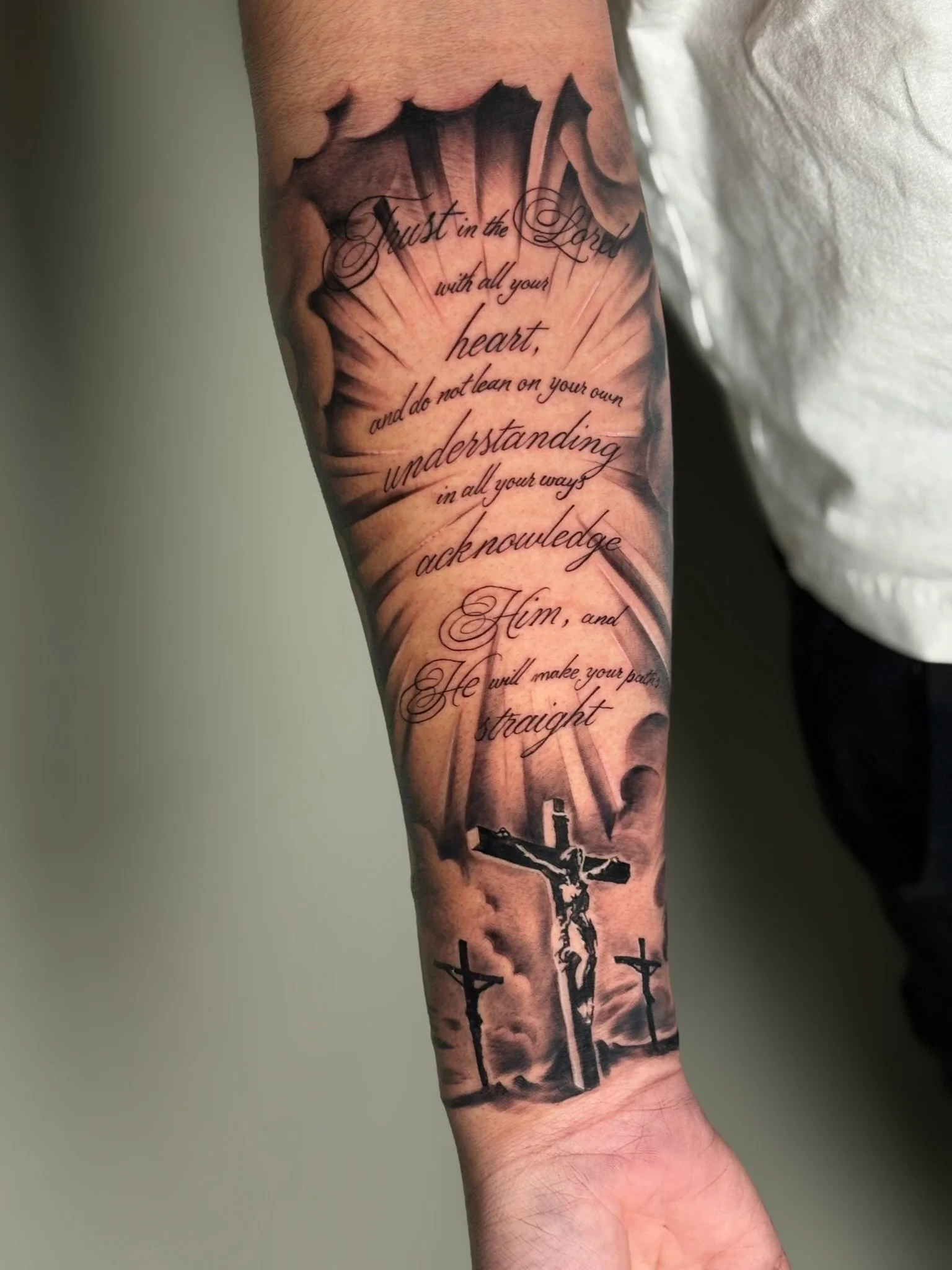 Tattoo of a crucifix with multiple crosses on the arm, along with a Bible verse written in cursive script.