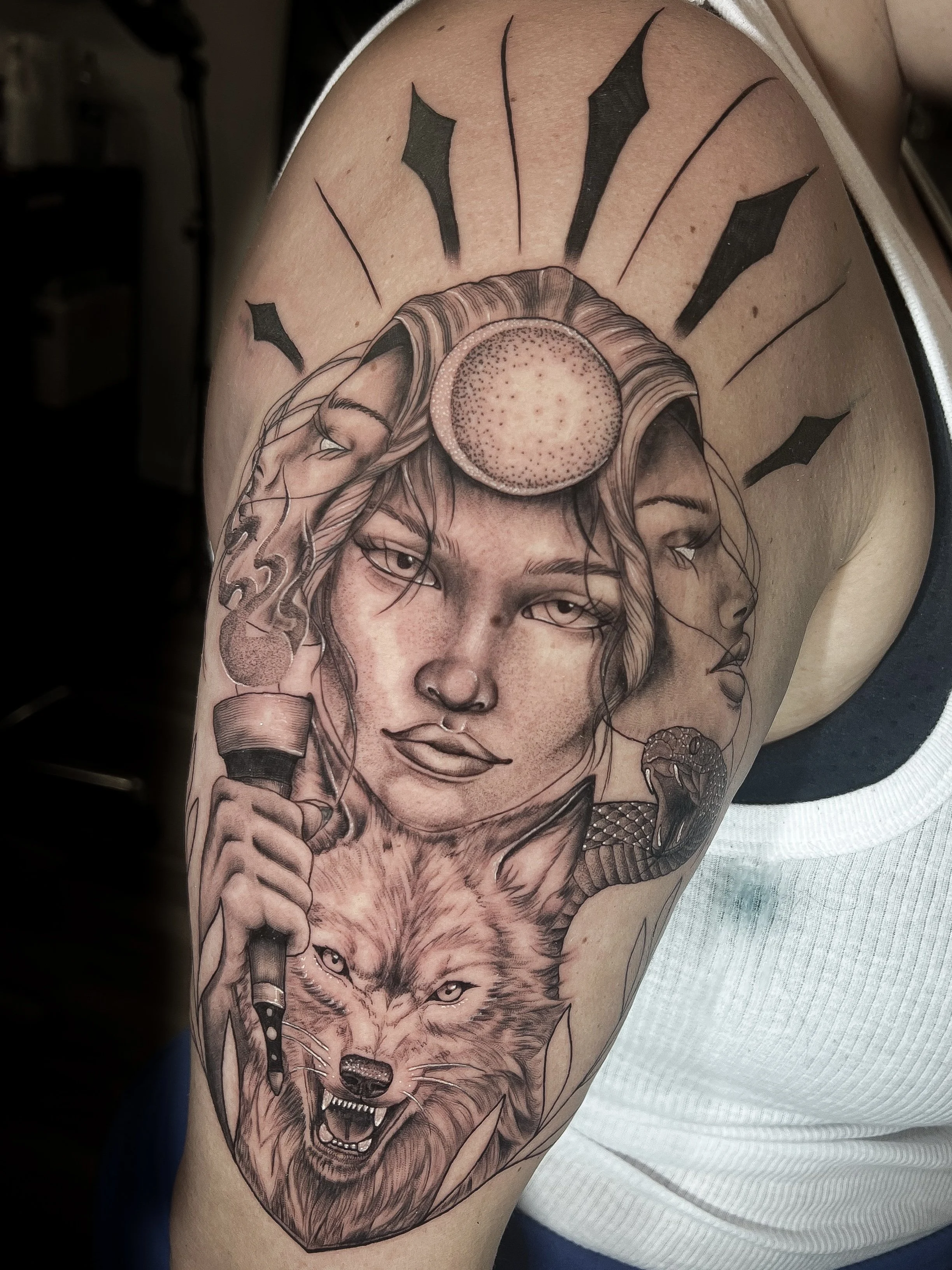 A detailed black and gray tattoo featuring a Goddess with long hair, holding a megaphone, a snarling wolf, and snakes on the upper arm.