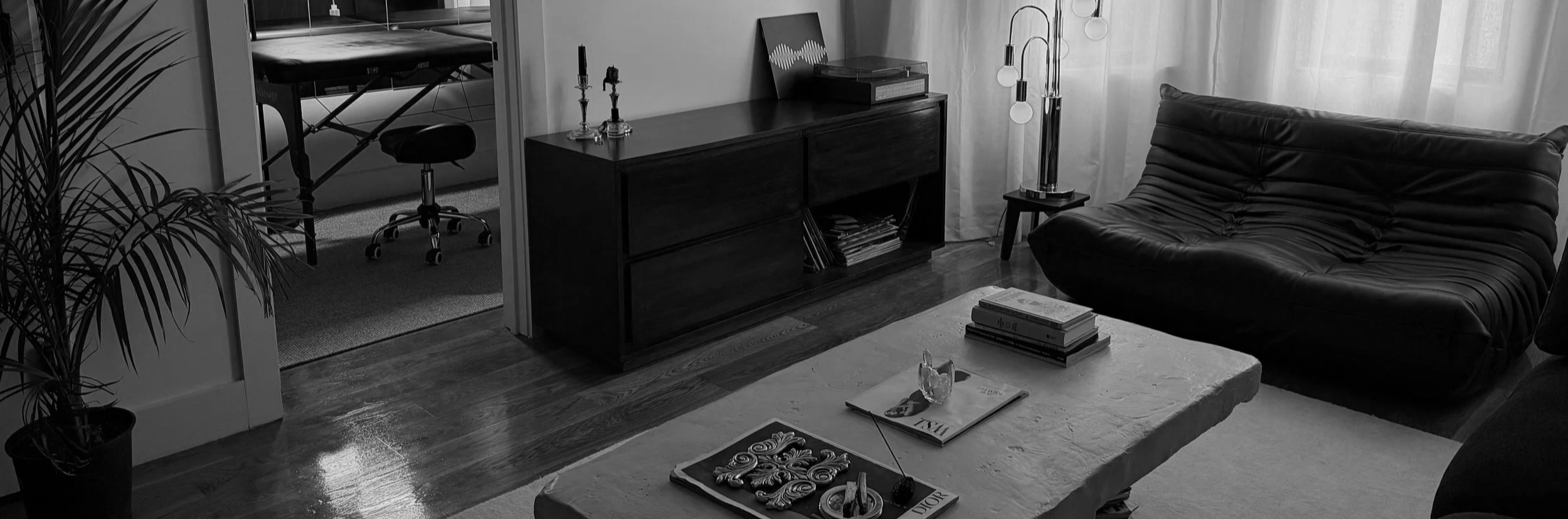 Living room with a black leather couch, a low coffee table with magazines and decorative items, a TV stand with record albums, and a floor lamp, with a window covered by curtains.