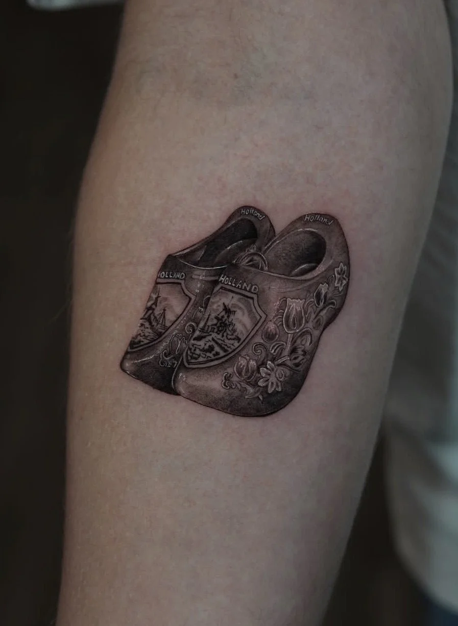 Tattoo of stacked Dutch clogs with designs and the word "HOLLAND" on each clog, placed on person's forearm.
