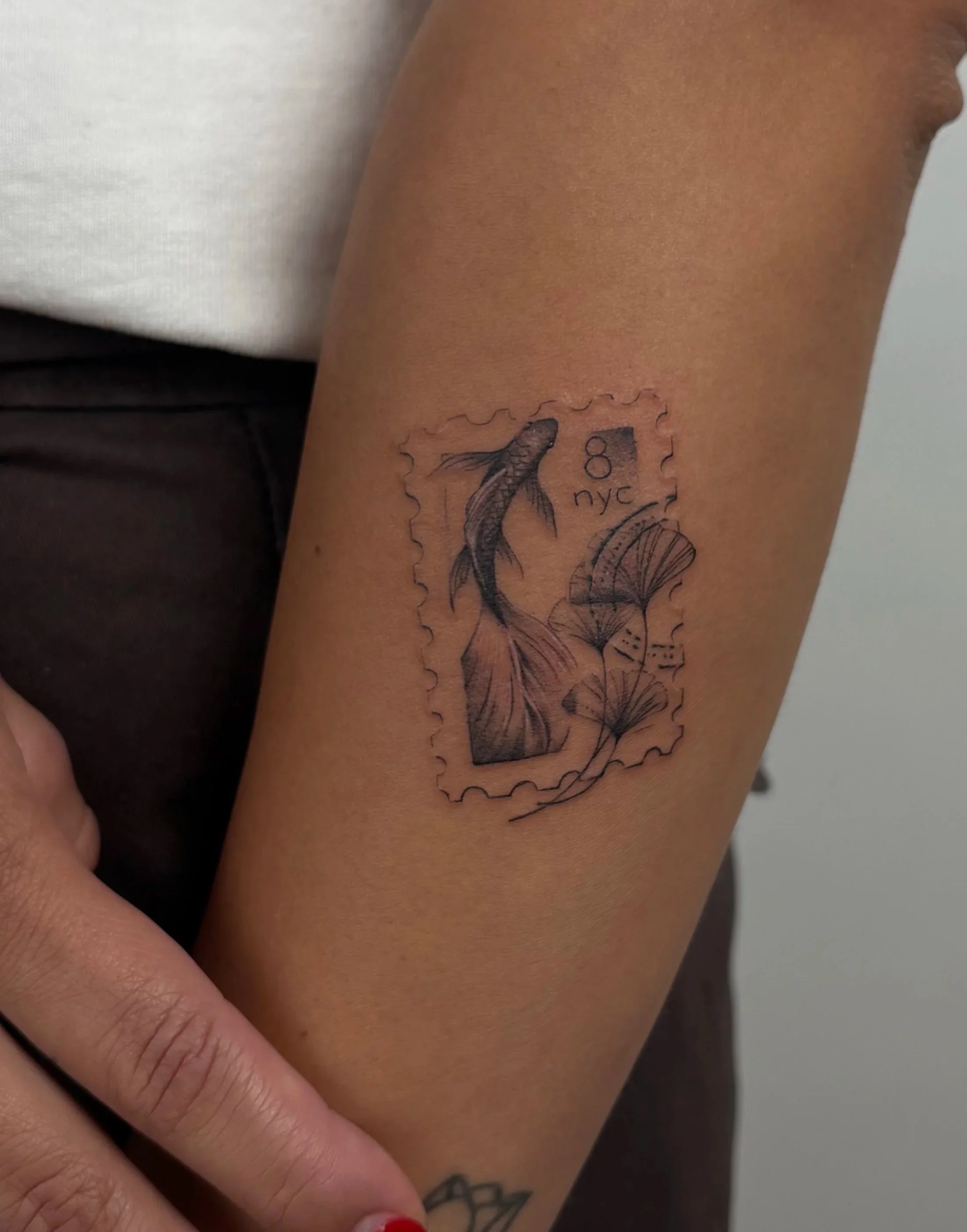 Close-up of a person's forearm with a black ink tattoo of a fish stamp, the letters 'NYC,' and a small drawing of a shell, surrounded by a stamped postage outline.