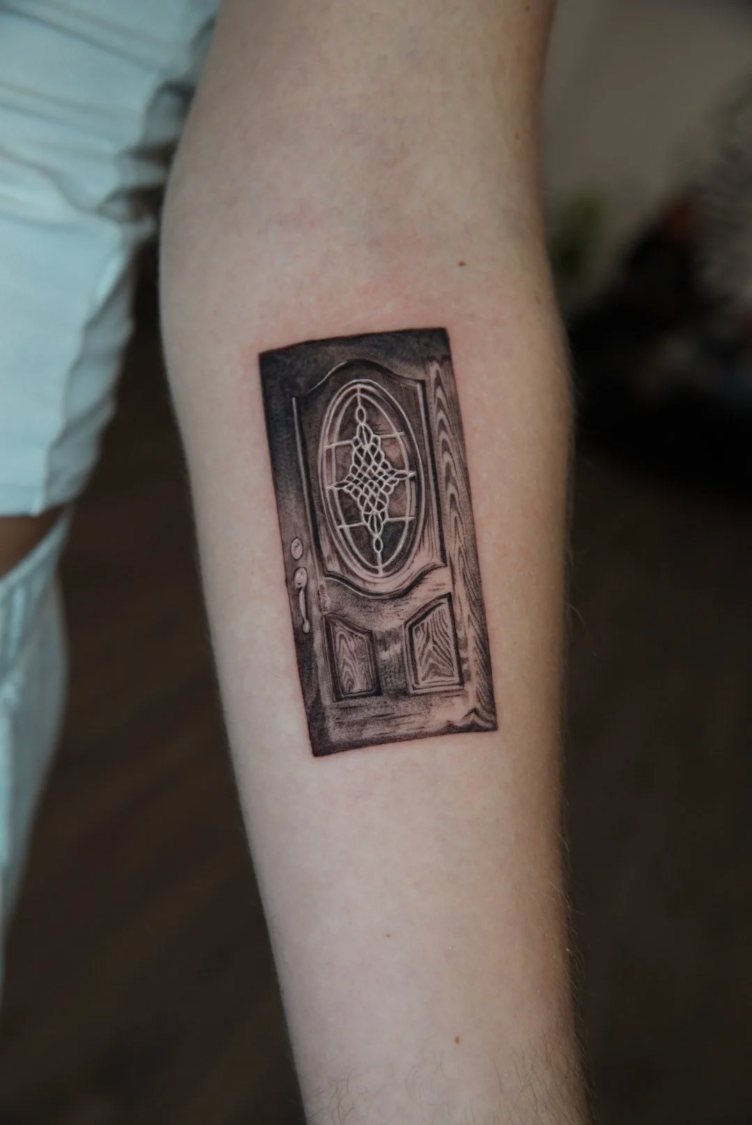 Tattoo of a detailed wooden door with a large oval window and spiderweb design on the glass, located on a person's arm.