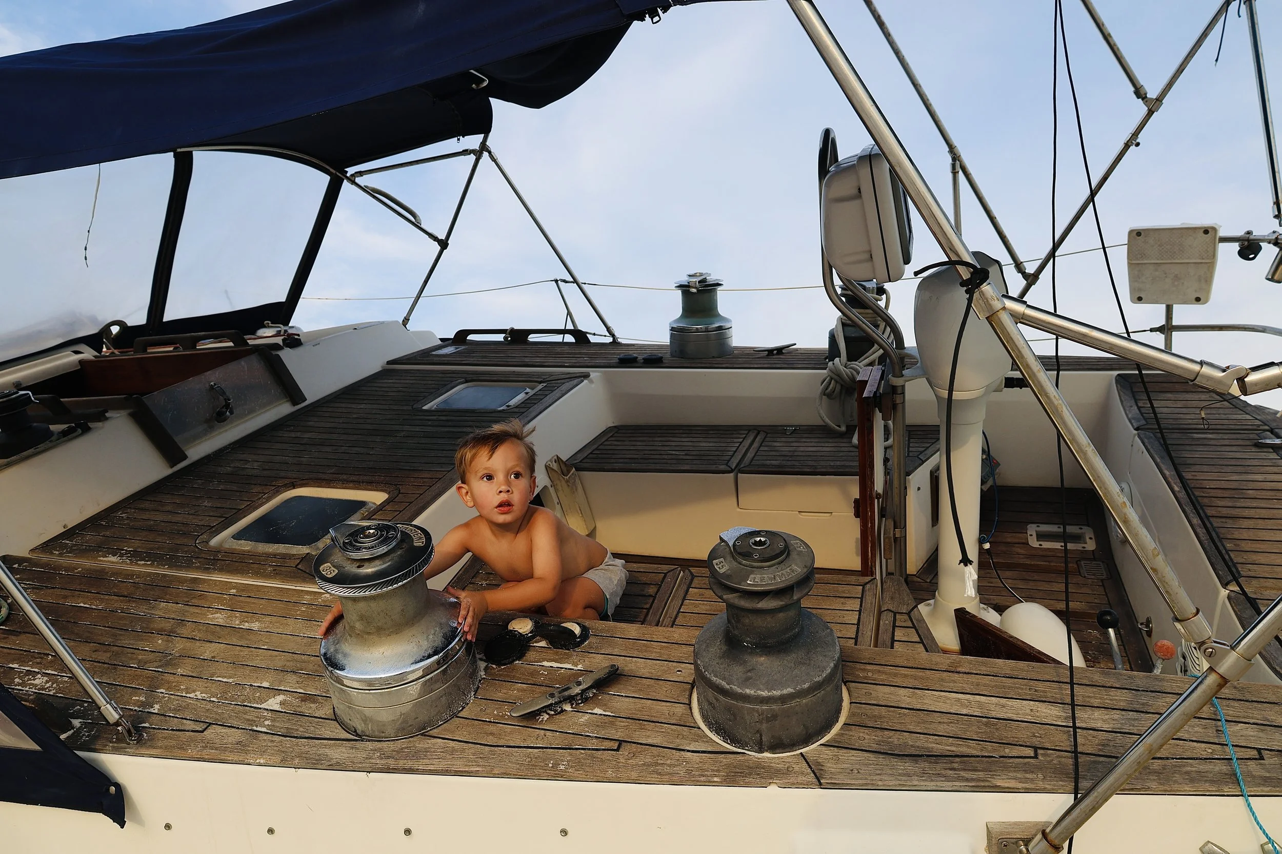 A young boy sitting on the wooden deck of a sailboat, surrounded by boat hardware and ropes, with the sky in the background.