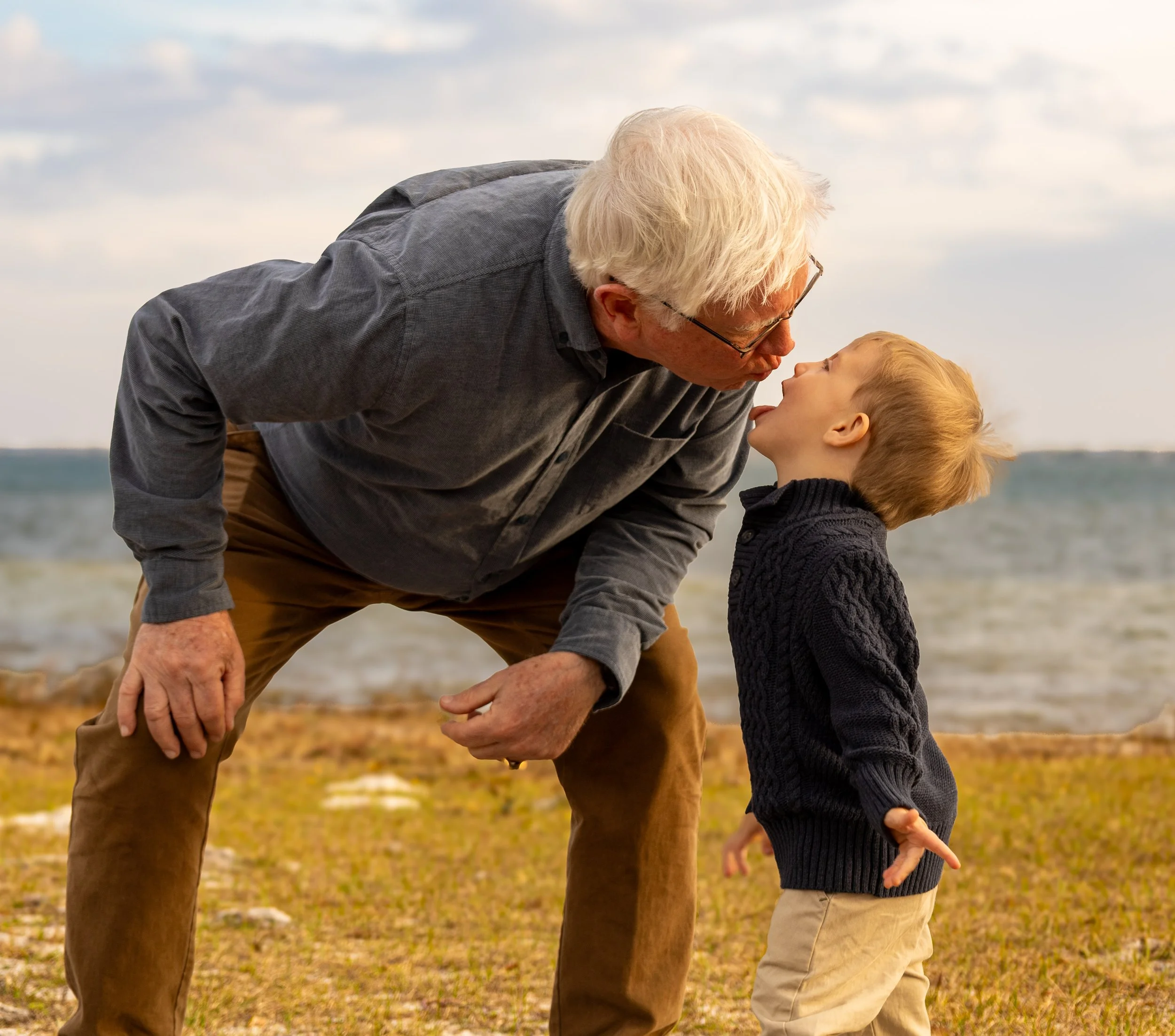 An elderly man with white hair and glasses leaning down to kiss a young boy with blond hair. They are outdoors near a body of water, with cloudy skies and grass in the background.