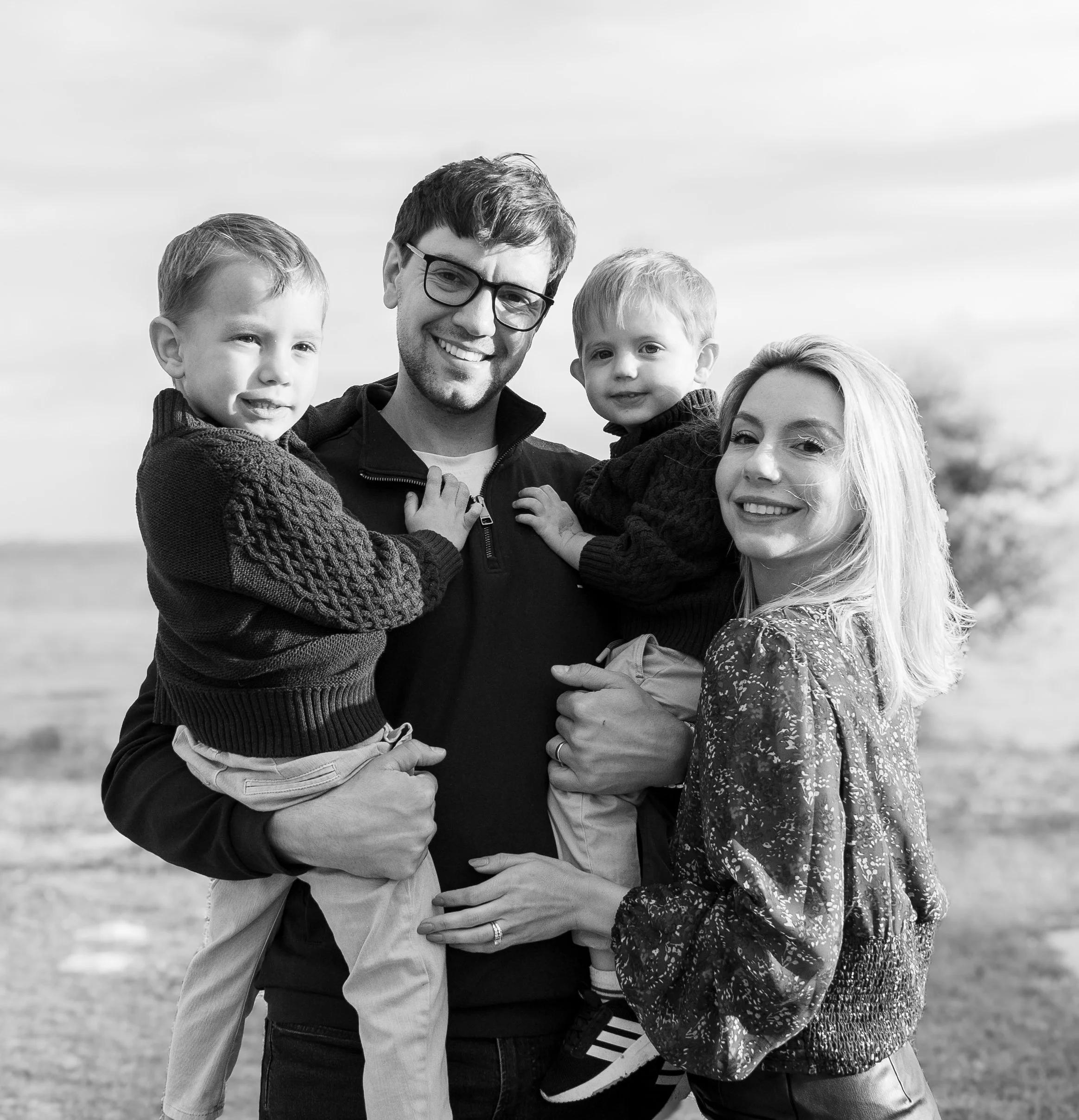 A family of four outdoors, smiling and standing close together. The father has dark hair, glasses, and a beard, and is holding two young boys, one on each side. The mother has long blonde hair and is smiling at the camera. The boys have light hair and are dressed warmly, one in a knitted sweater and the other in a jacket. The background shows a cloudy sky, and the photo is in black and white.