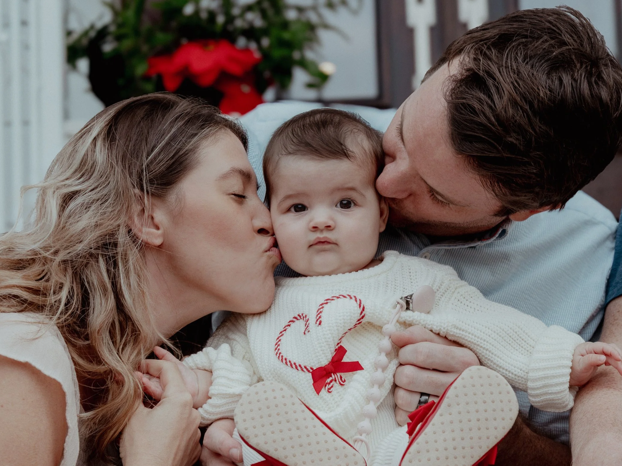 A family of three: a mother, father, and baby, sharing a tender moment. The mother and father kiss the baby's cheeks, who has wide eyes and looks directly at the camera. The background features a poinsettia plant.