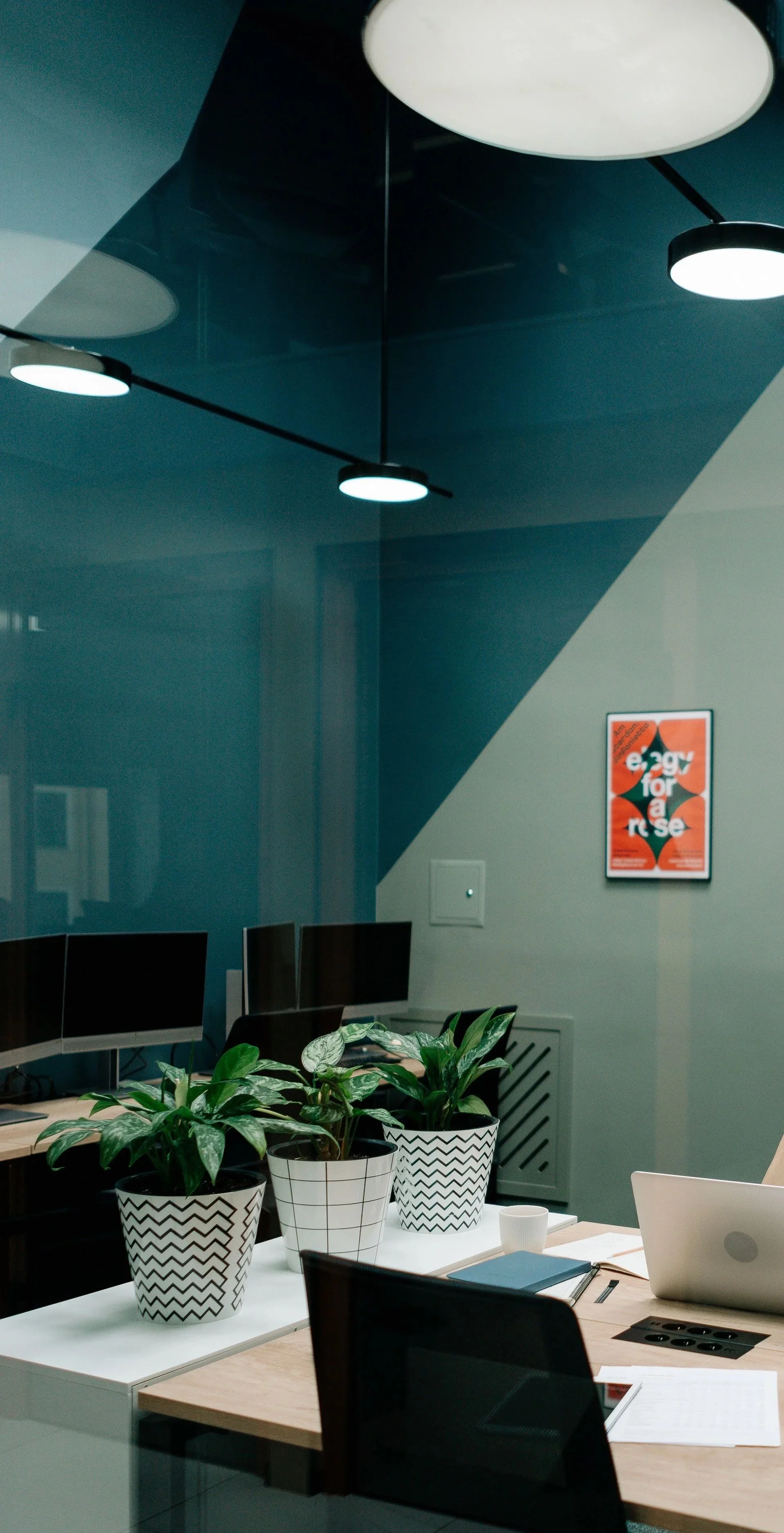 Office meeting room with a conference table, laptops, notebooks, a cup, potted plants, a wall poster, and ceiling lights.
