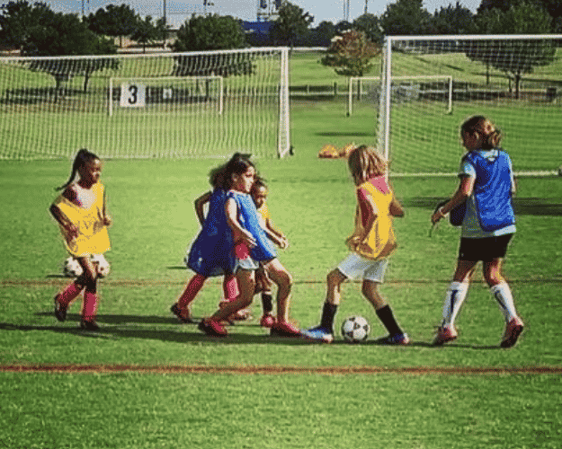 Young girls playing soccer on a grass field, some wearing yellow jerseys and others in blue jerseys, with a soccer ball at the center and a goalpost in the background.