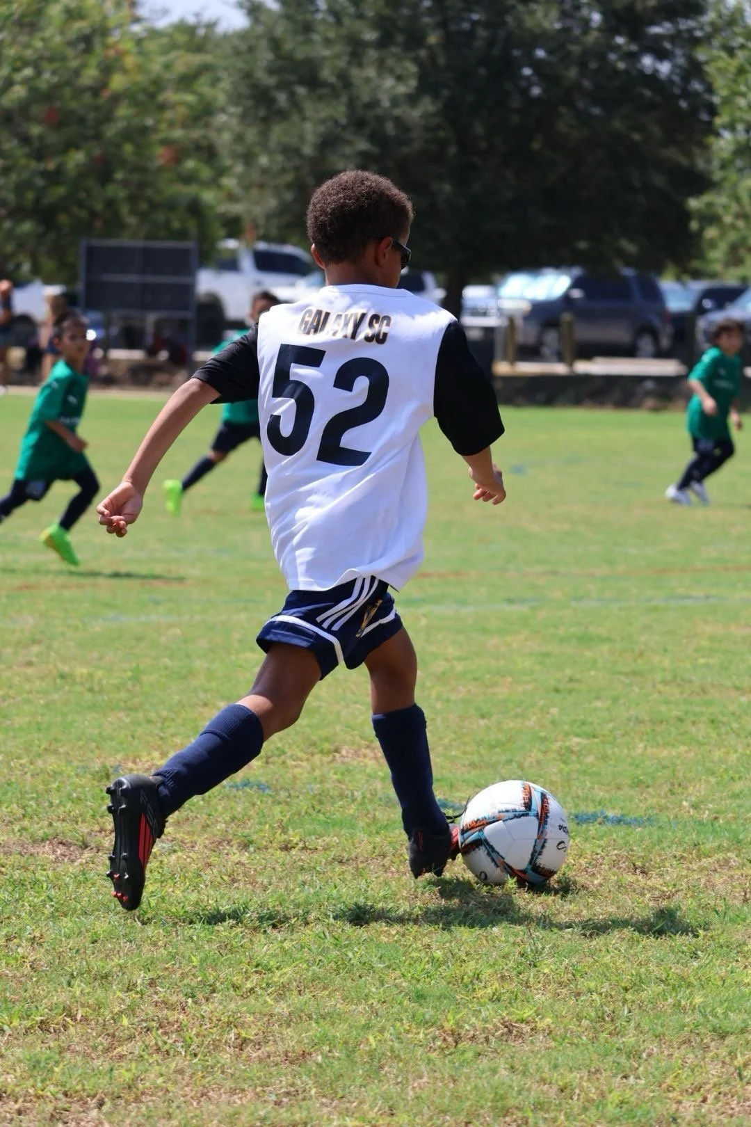 A young boy playing soccer on a grassy field, wearing a white jersey with the number 52, black shorts, and black cleats, kicking a soccer ball. Other children are playing in the background.