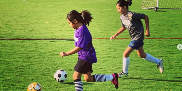 Two young girls playing soccer on a green field, running after the ball, with a soccer goal in the background.