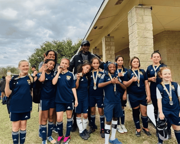 Group of young female soccer players and their coach outdoors, wearing blue uniforms and medals.