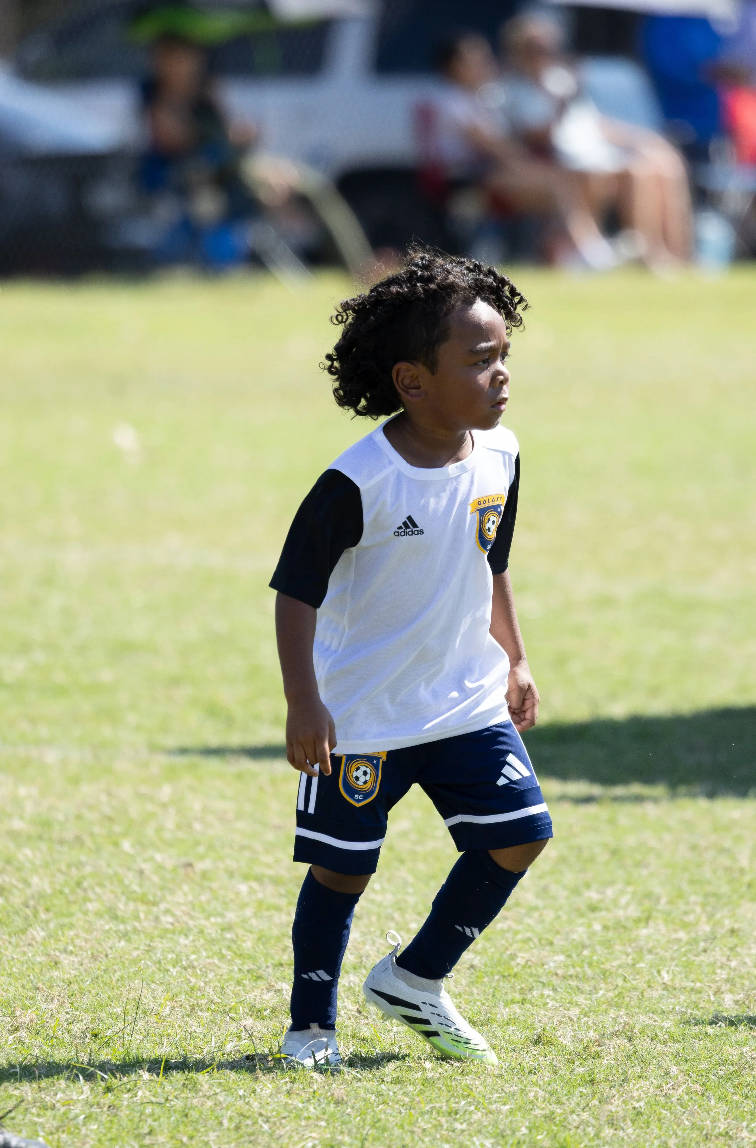 Young boy with curly hair wearing a soccer uniform standing on a grassy field during a soccer game.
