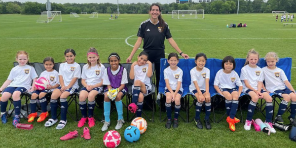 Group of young girls in soccer uniforms sitting on a bench with their coach on a soccer field.