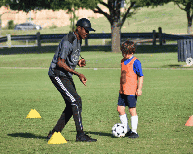 A man coaching a young boy on how to play soccer on a grassy field, with cones set up for drills and a tree in the background.