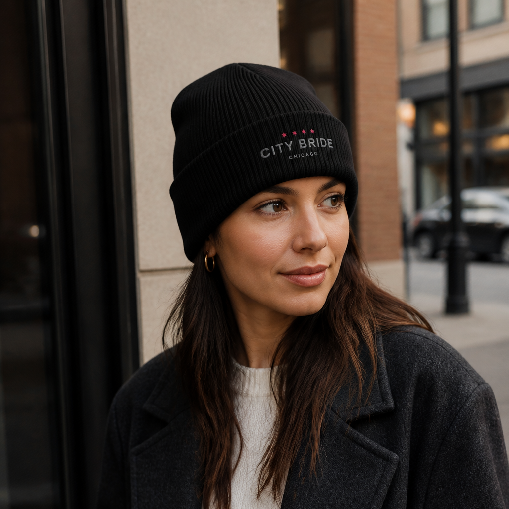 A woman wearing a black knit beanie with 'City Bride Chicago' embroidered on it, dressed in a dark coat and white shirt, stands outdoors in front of a glass window and brick building in an urban setting.