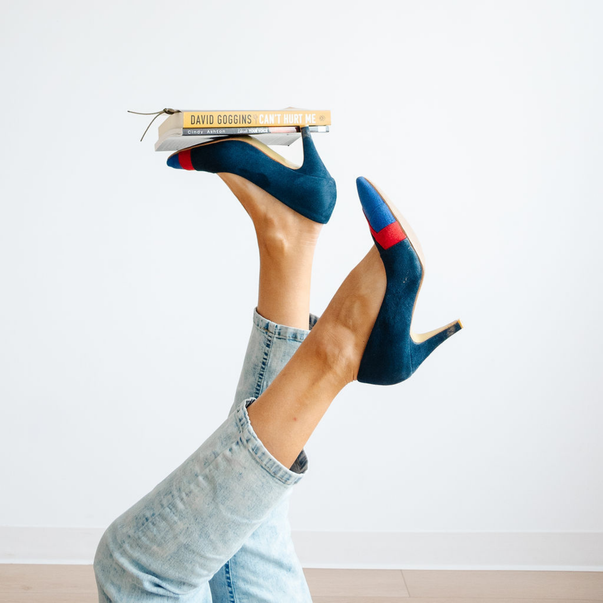 Person in blue high-heeled shoes with red accents, wearing light blue jeans, holding a stack of books against a plain white wall.