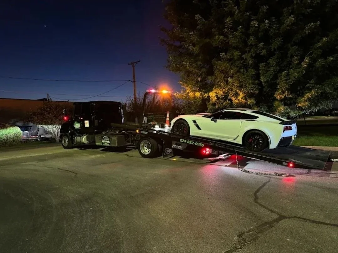 White sports car on a tow truck at night with street and trees in the background.