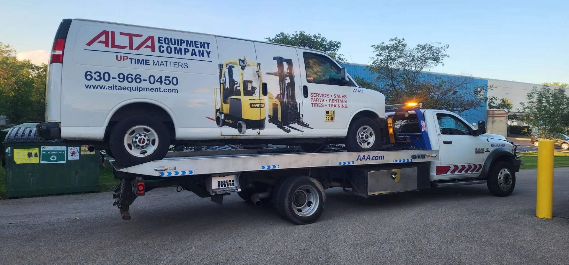 A tow truck carrying a white van with advertisements for Alta Equipment Company, showing forklift equipment and contact information, parked outdoors near trees and a building.