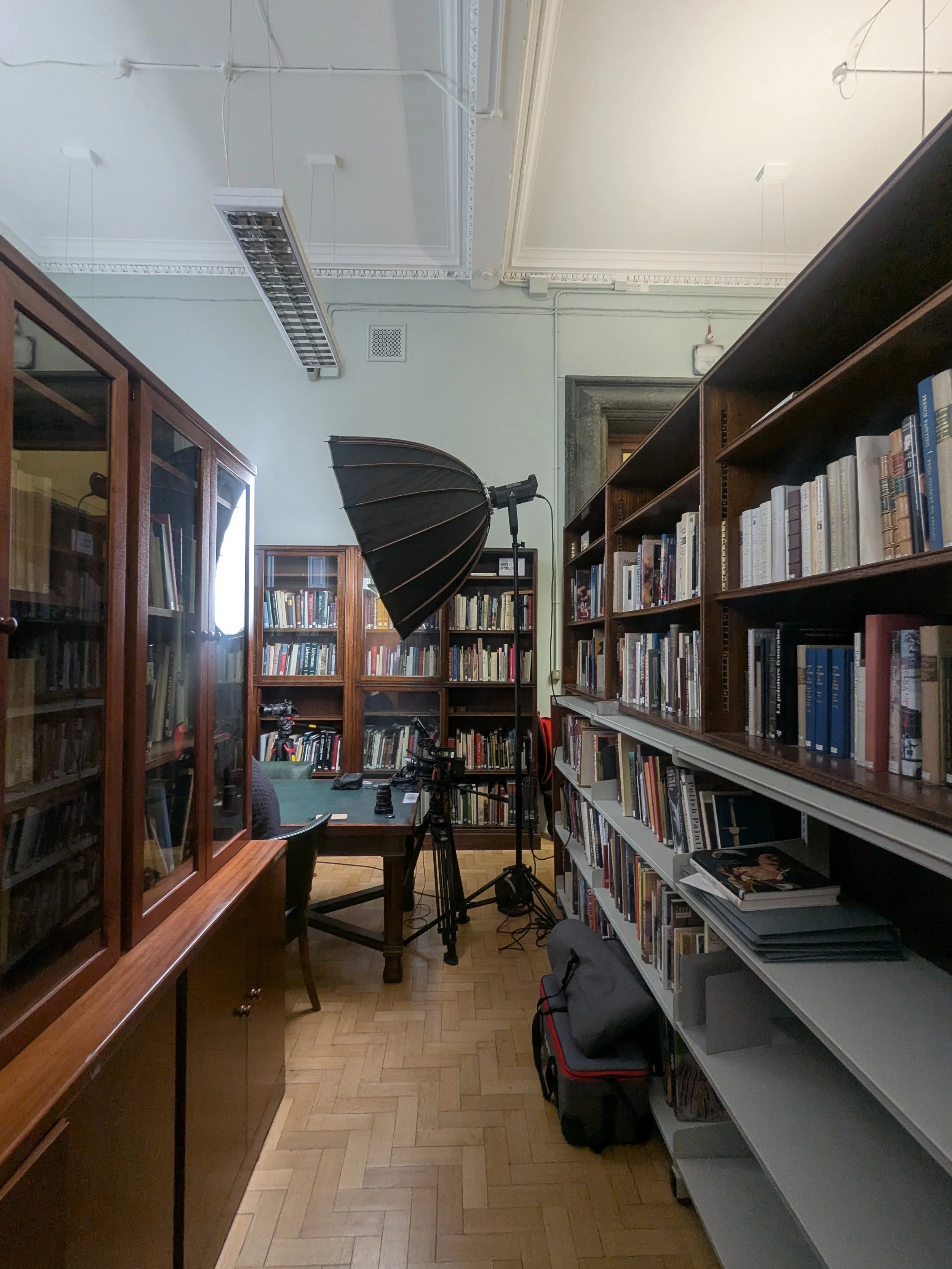 Photo of a room with wooden bookshelves filled with books on the right and a wooden cabinet with glass doors filled with books on the left. In the center, there is a table with a camera on a tripod and some electronic equipment. A large softbox light is set up in front of the table, aimed at the bookshelves. The room has a parquet wood floor and a high ceiling with decorative molding.