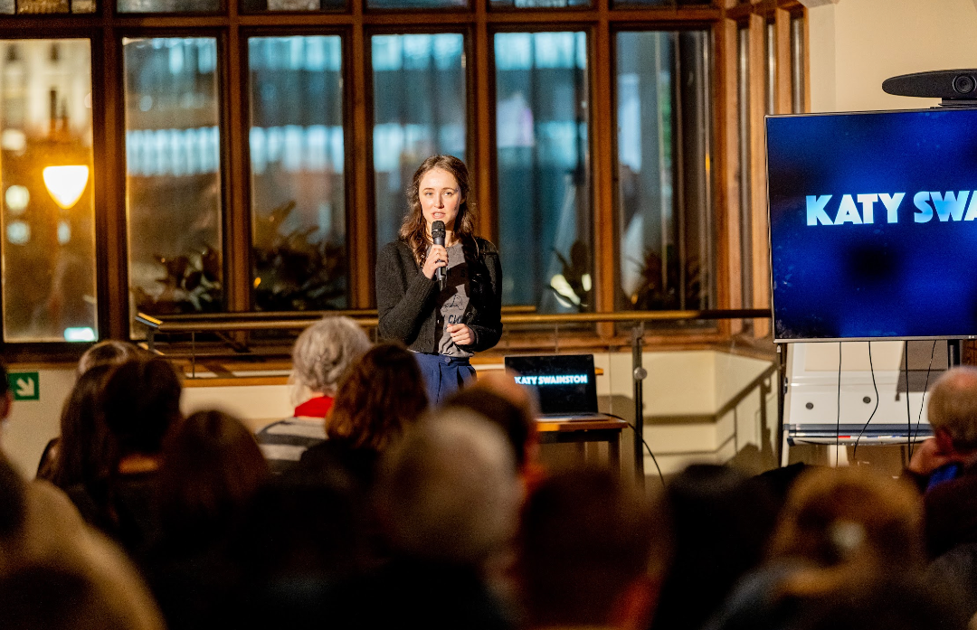 A woman with long brown hair is speaking into a microphone. She is standing in front of a large window with wooden frames, and there is a large screen displaying the name 'Katy Swainston' to her right. The audience is seated in front of her.