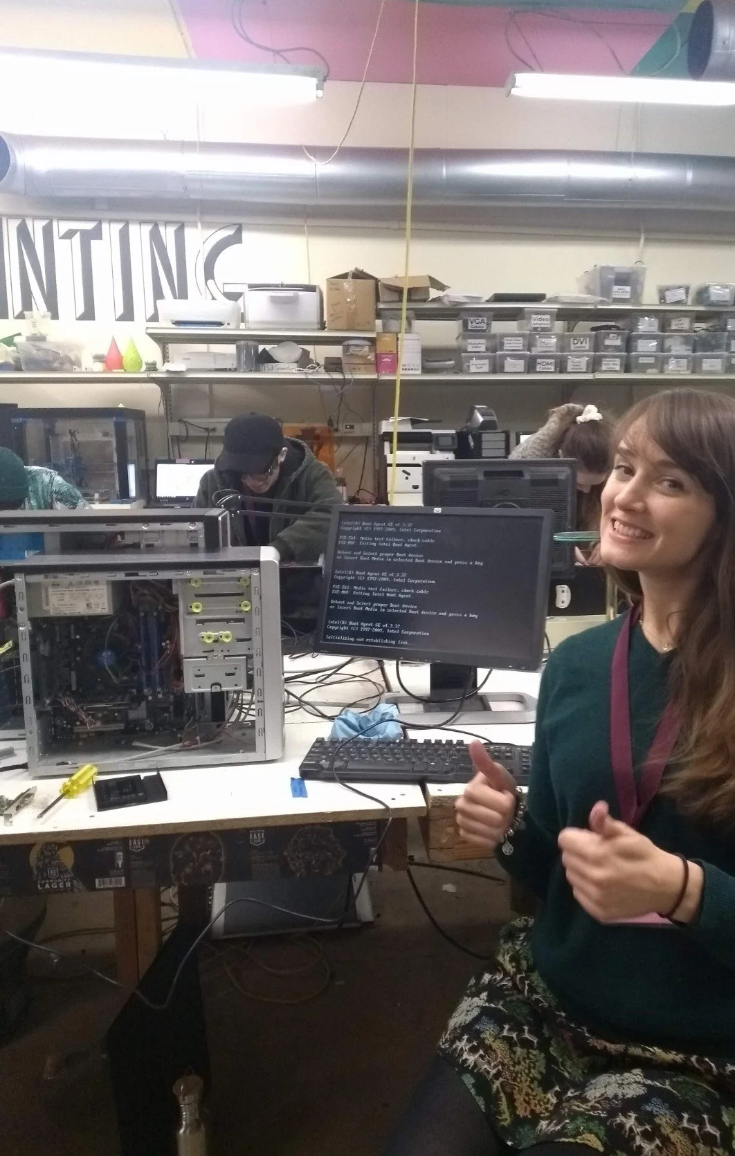 A woman giving a thumbs up in a busy electronics workshop with a broken desktop computer on her desk, people working in the background, and computer components and tools visible.