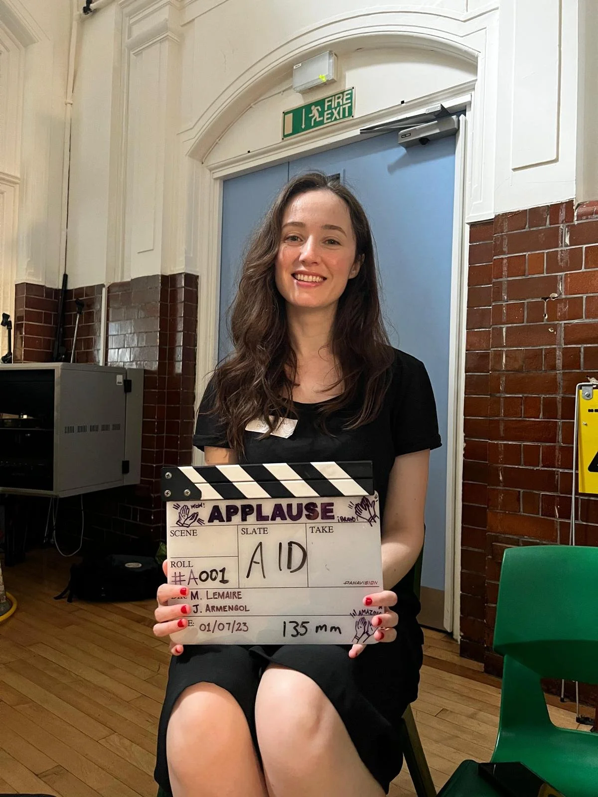 Katy Swainston, a woman with brown hair is holding a clapperboard with 'APPLAUSE' written on it, seated indoors against a brick wall background, smiling, with a blue door and fire exit sign behind her.