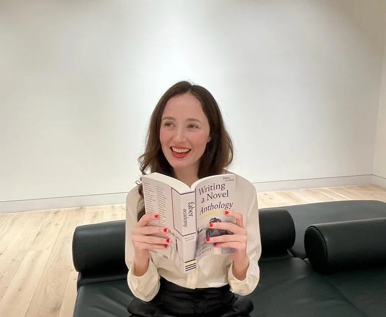 Katy Swainton, a woman with long brown hair, wearing a white blouse, is sitting on a black leather couch in a minimalist room with wooden floors and a white wall. She is smiling and holding an open book titled 'Writing a Novel Anthology.'