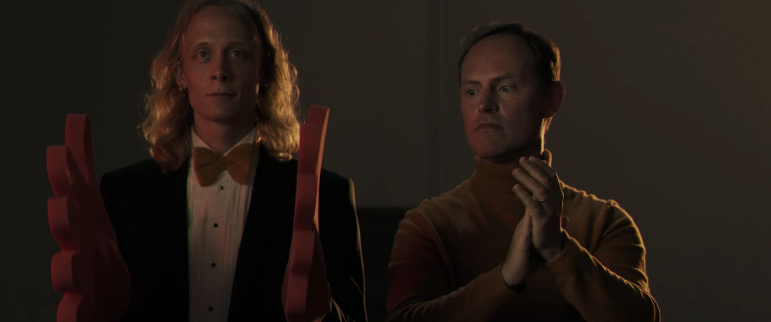 A scene from a film called Applause showing two men, one with long blonde hair with a bow tie, and the other with short hair wearing a brown turtleneck, in a dark room, applauding. The man with the long hair is hear huge, red foam hands.