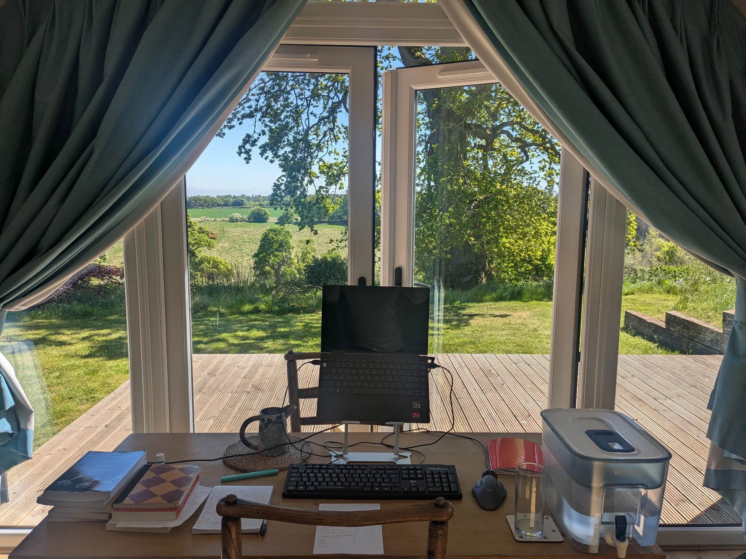 An indoor workspace with a wooden desk and a large window overlooking a green landscape with trees and fields. The desk has a computer monitor, a wireless keyboard, a mouse, notebooks, a coffee mug, a water dispenser, and some papers. Blue curtains are pulled back on both sides of the window.