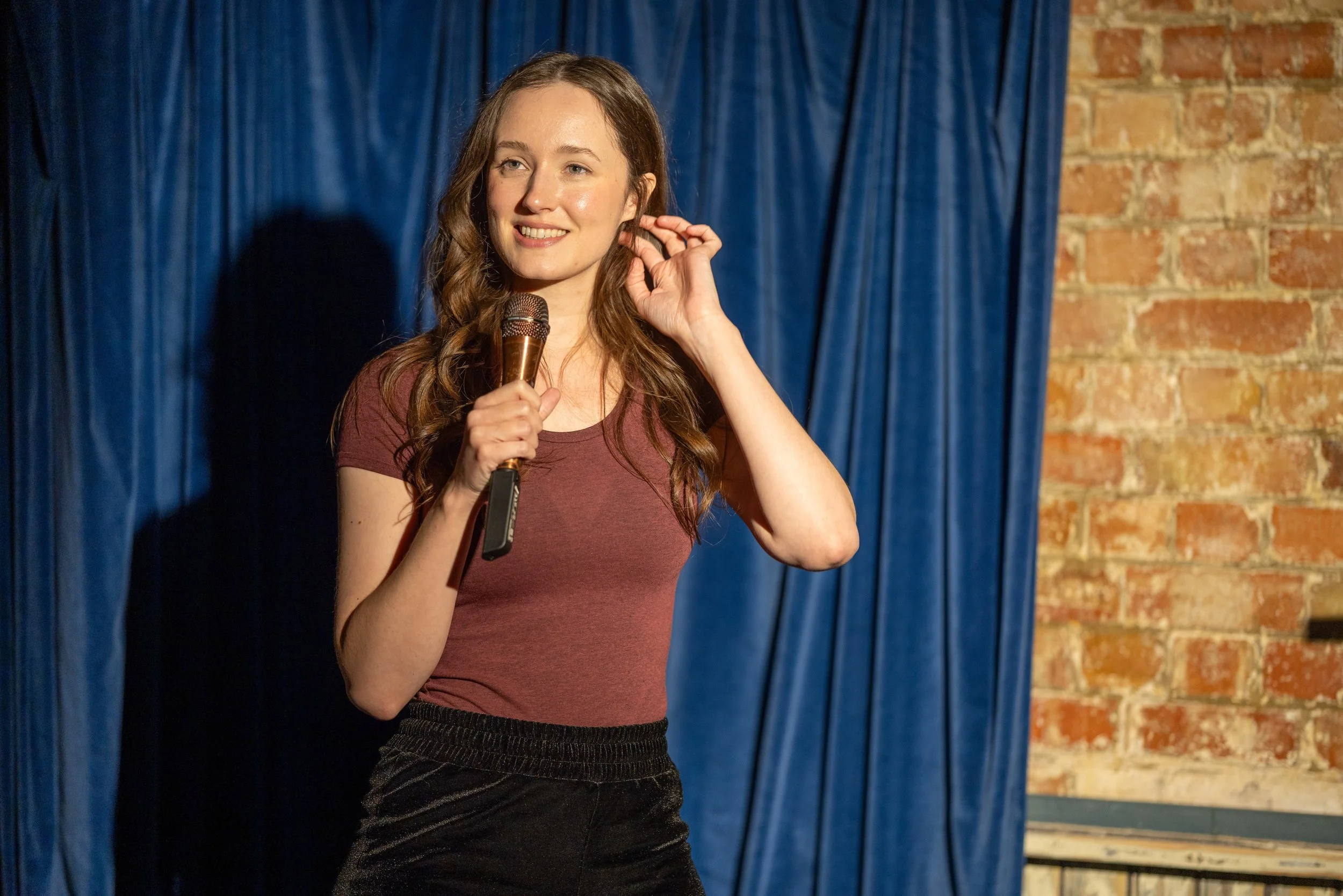 Katy Swainston, a woman with long, wavy brown hair, is holding a microphone and smiling, standing in front of a blue curtain and a brick wall while performing stand-up.