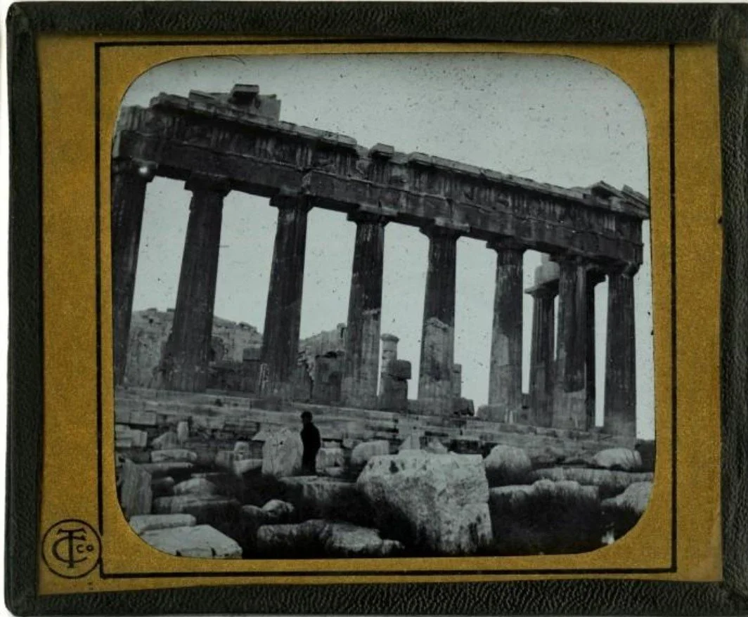 Black and white photo of ancient ruins, including the Parthenon temple with tall columns, surrounded by scattered rocks and a person standing among the ruins.