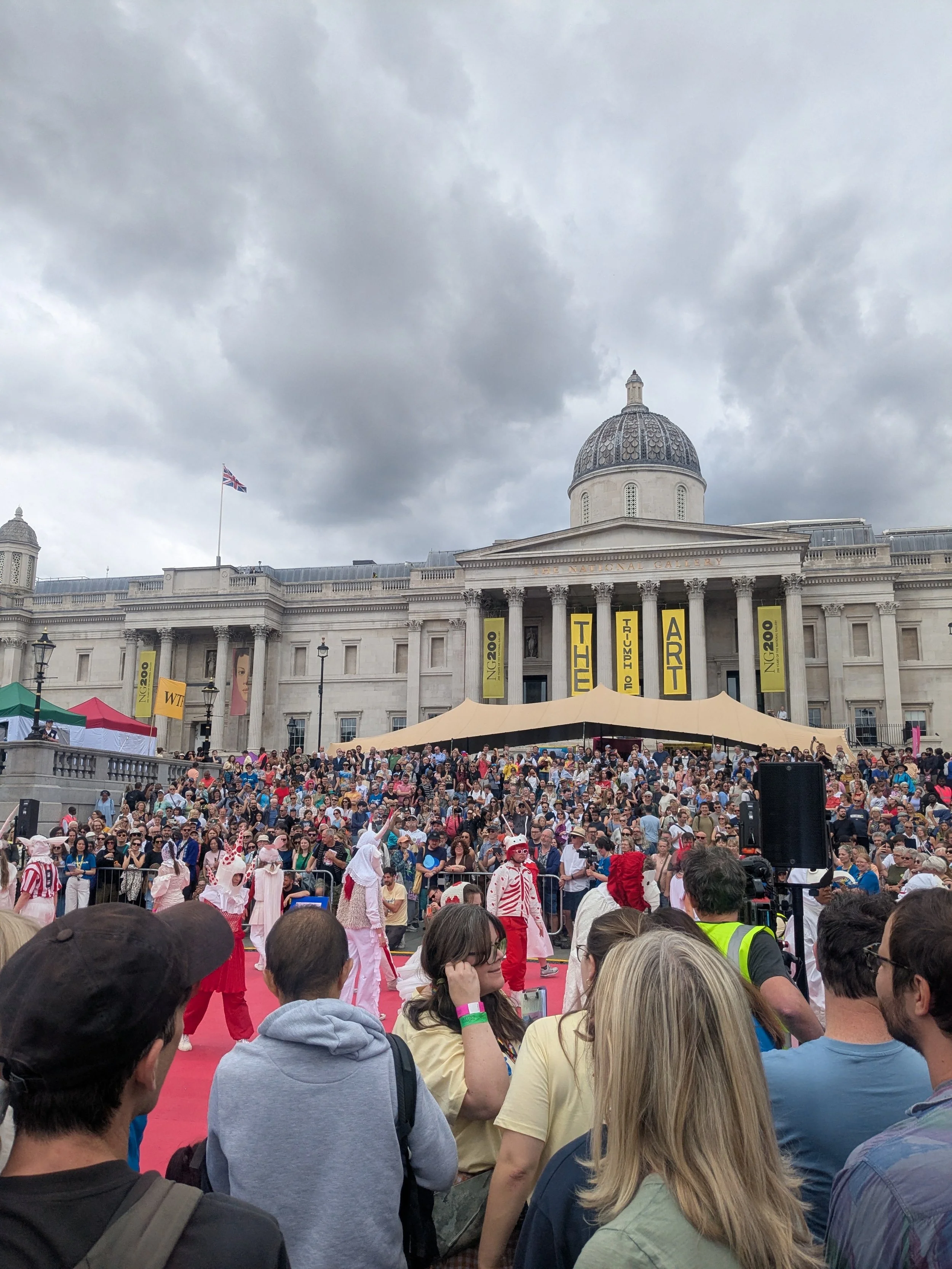 Crowd gathered in front of a large building with a domed roof and columns, under a cloudy sky, during a public event or gathering. The building is the National Gallery and the event is Jeremy Deller's The Triumph of Art.
